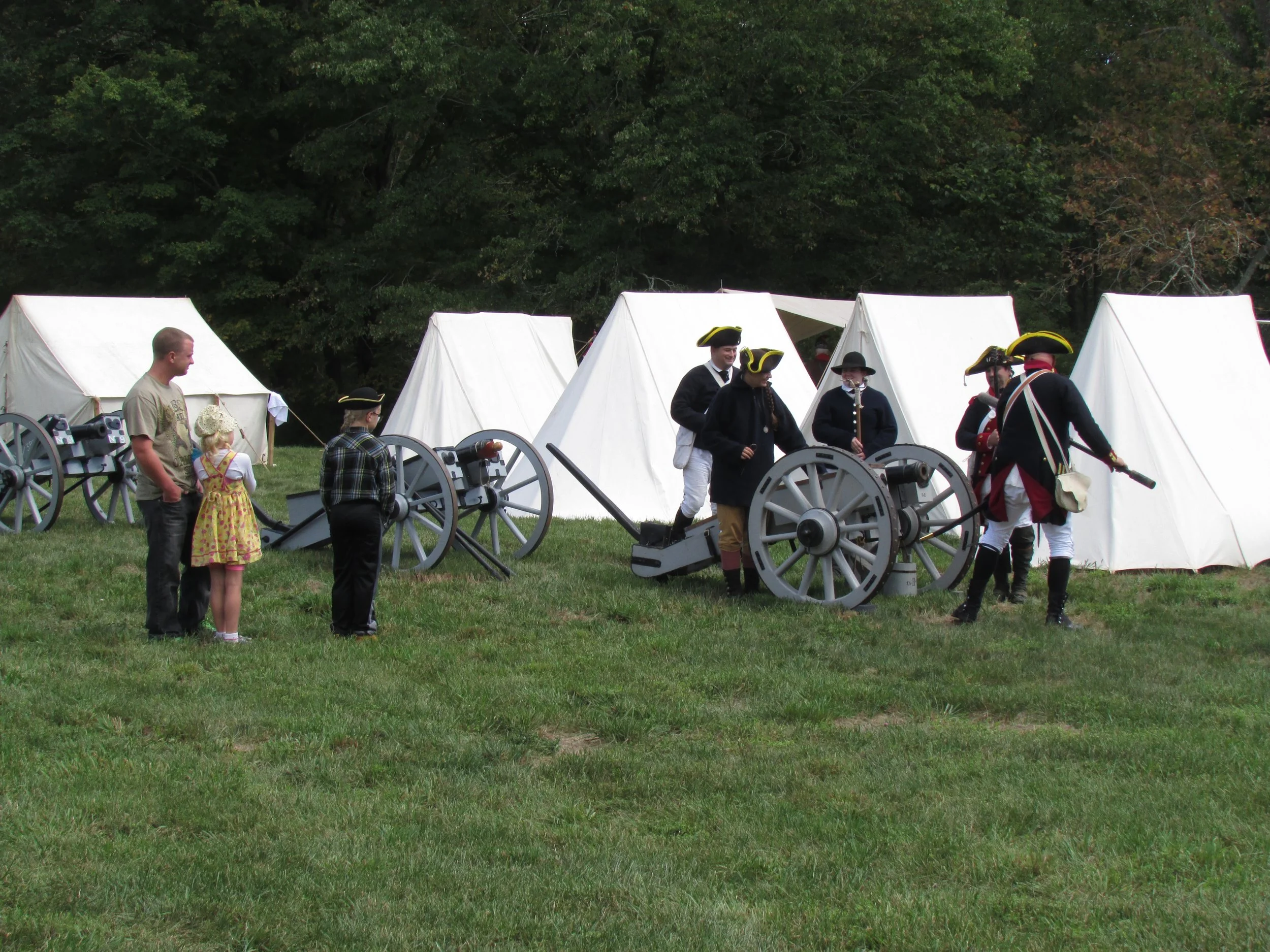 People dressed in historical military costumes from the American Revolutionary War era are participating in a reenactment on a grassy field, with white tents in the background.