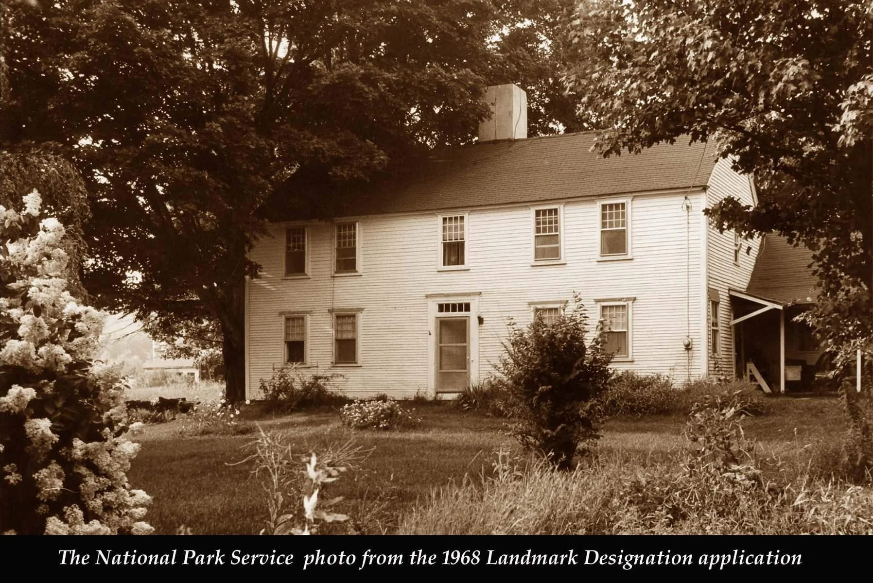 A sepia-toned photo of a two-story white wooden house with multiple windows and a sloped roof, surrounded by large trees and bushes, with a grassy yard in the foreground.