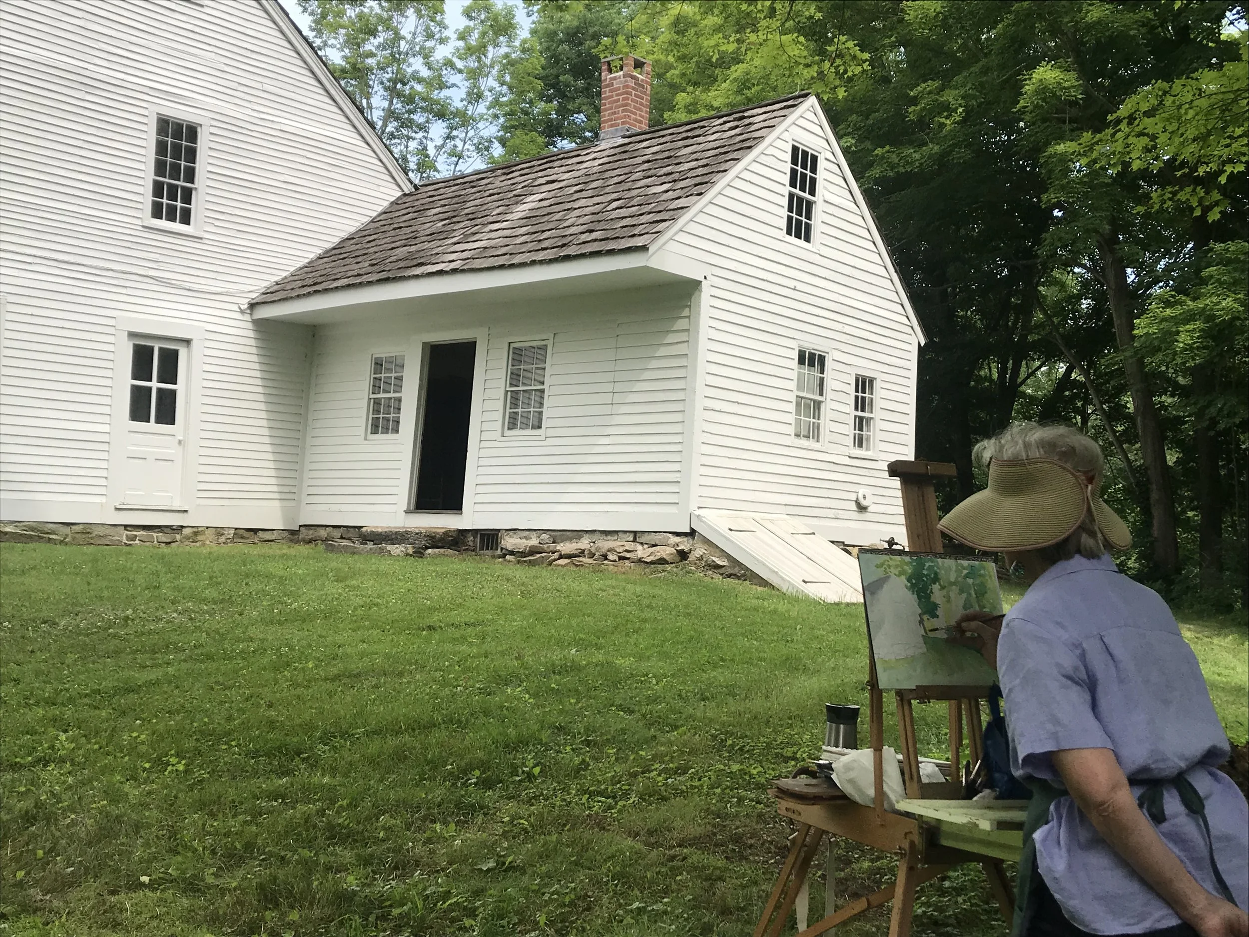 An artist painting a house on an easel in a grassy yard, with trees in the background.