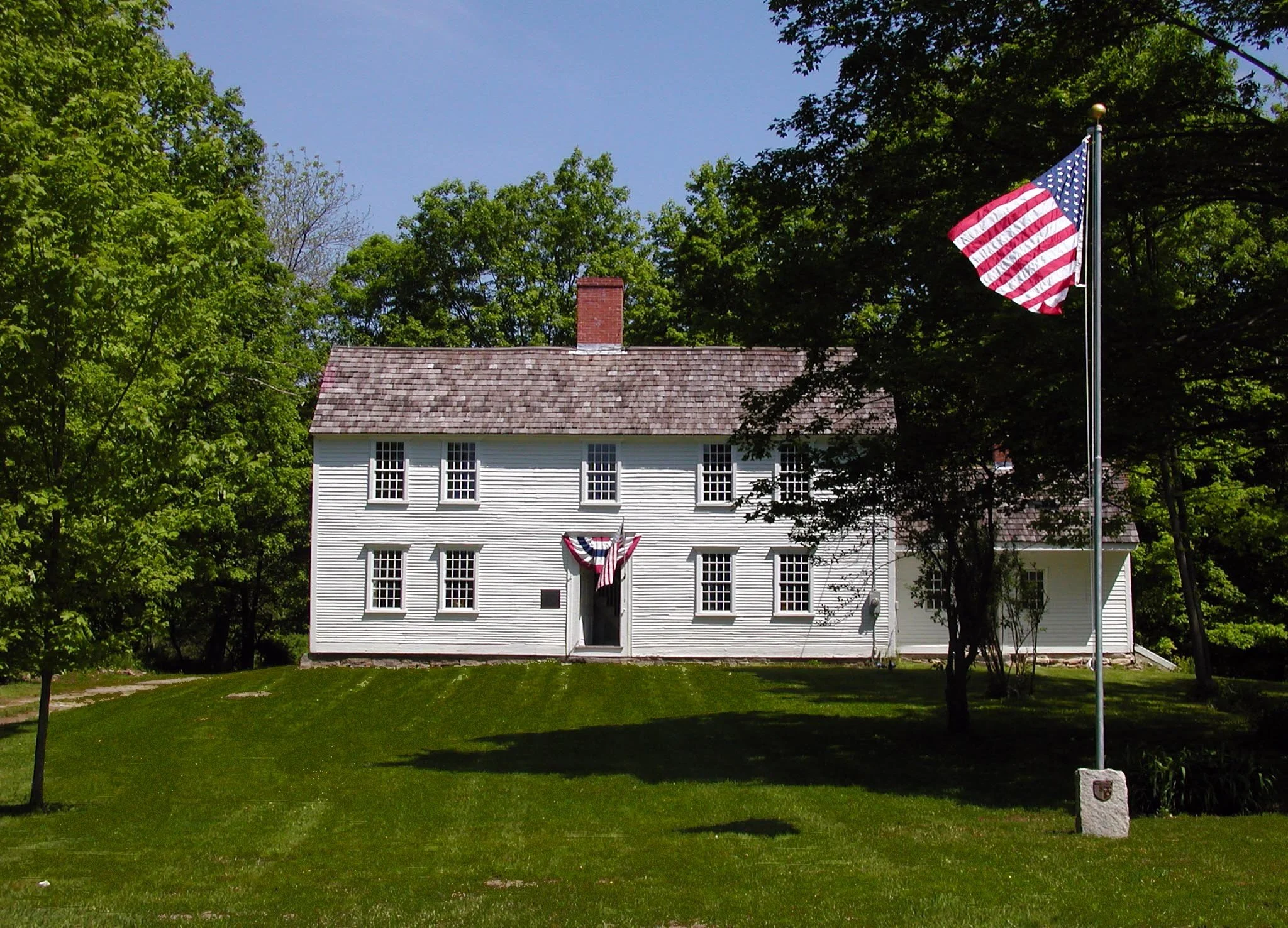 A white two-story house with a shingled roof surrounded by green trees and grass. An American flag is flying on a tall flagpole in front of the house, and a smaller American flag is draped over the front door. The house has six windows on the front facade, with some decorated with patriotic bunting.