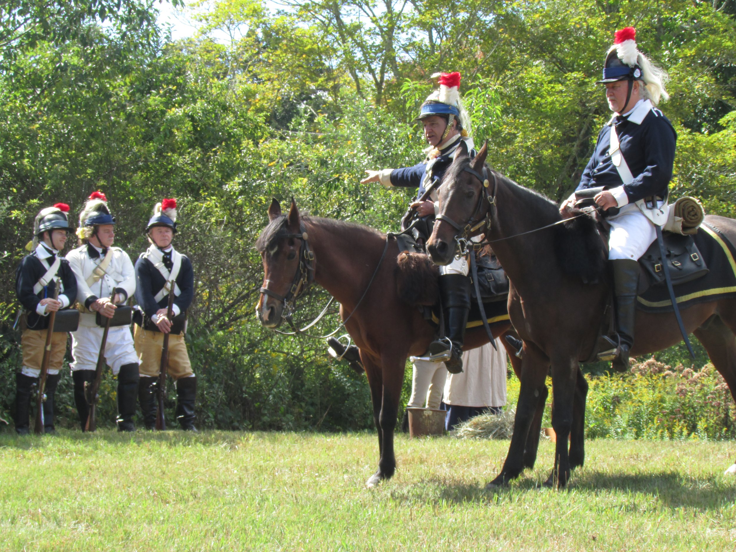 Historical reenactors dressed as soldiers from the American Revolutionary War, with some on horseback and others standing on a grassy field with trees in the background.