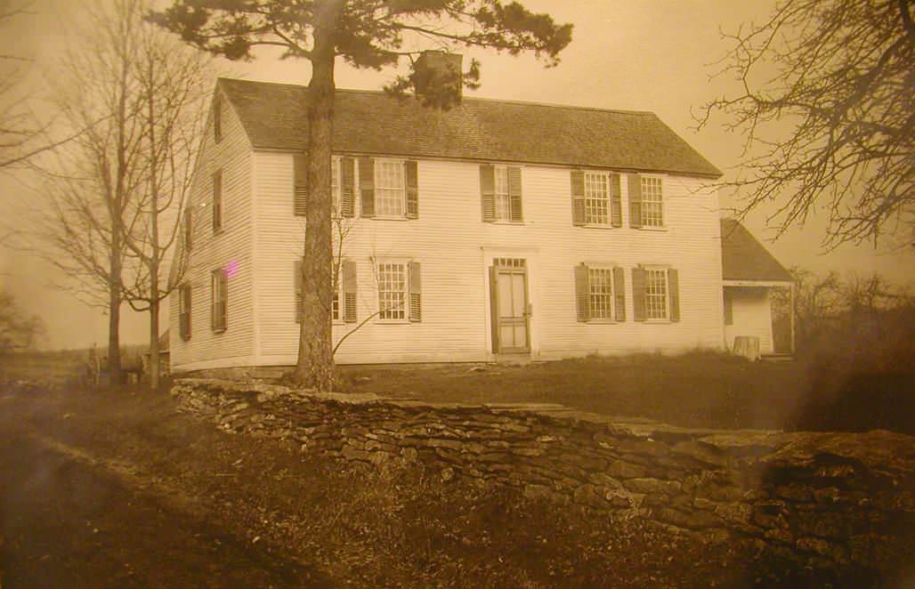 A large, historic two-story house with shuttered windows and a central front door, surrounded by leafless trees and a stone wall in the foreground, in sepia tone.