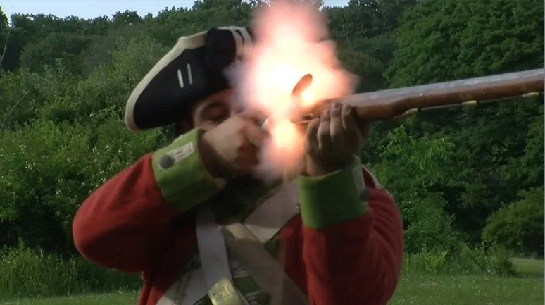 A person dressed in a historical uniform is firing a musket with a visible flame and smoke, outdoors in a green, wooded area.