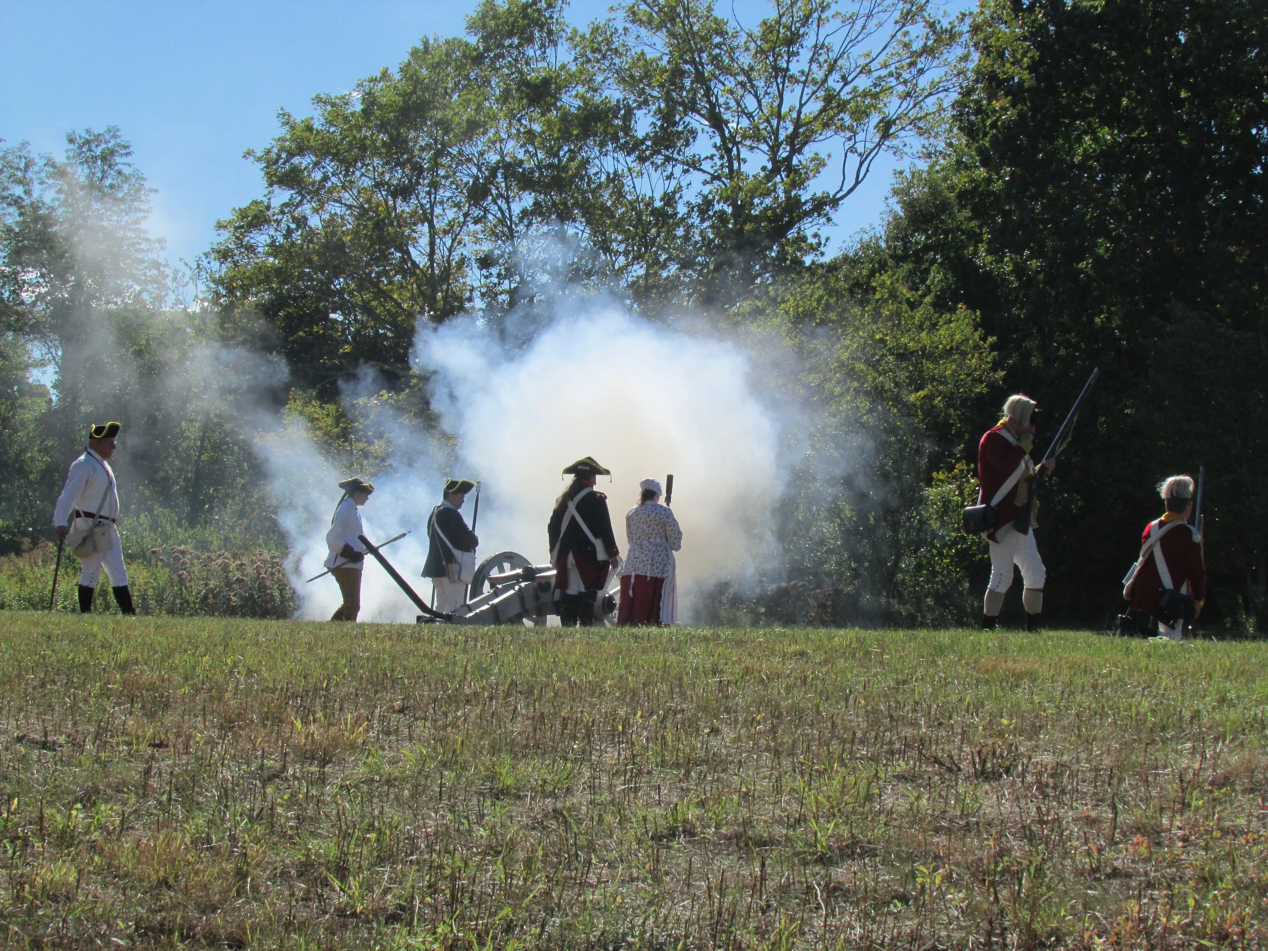 A group of people dressed in colonial military uniforms operating a cannon during a historical reenactment in a grassy field with trees in the background.