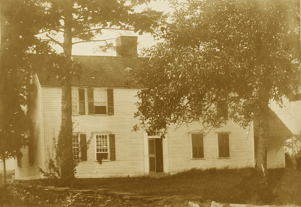 An old sepia-toned photograph of a two-story wooden house with multiple windows and shutters, surrounded by trees and a grassy yard.