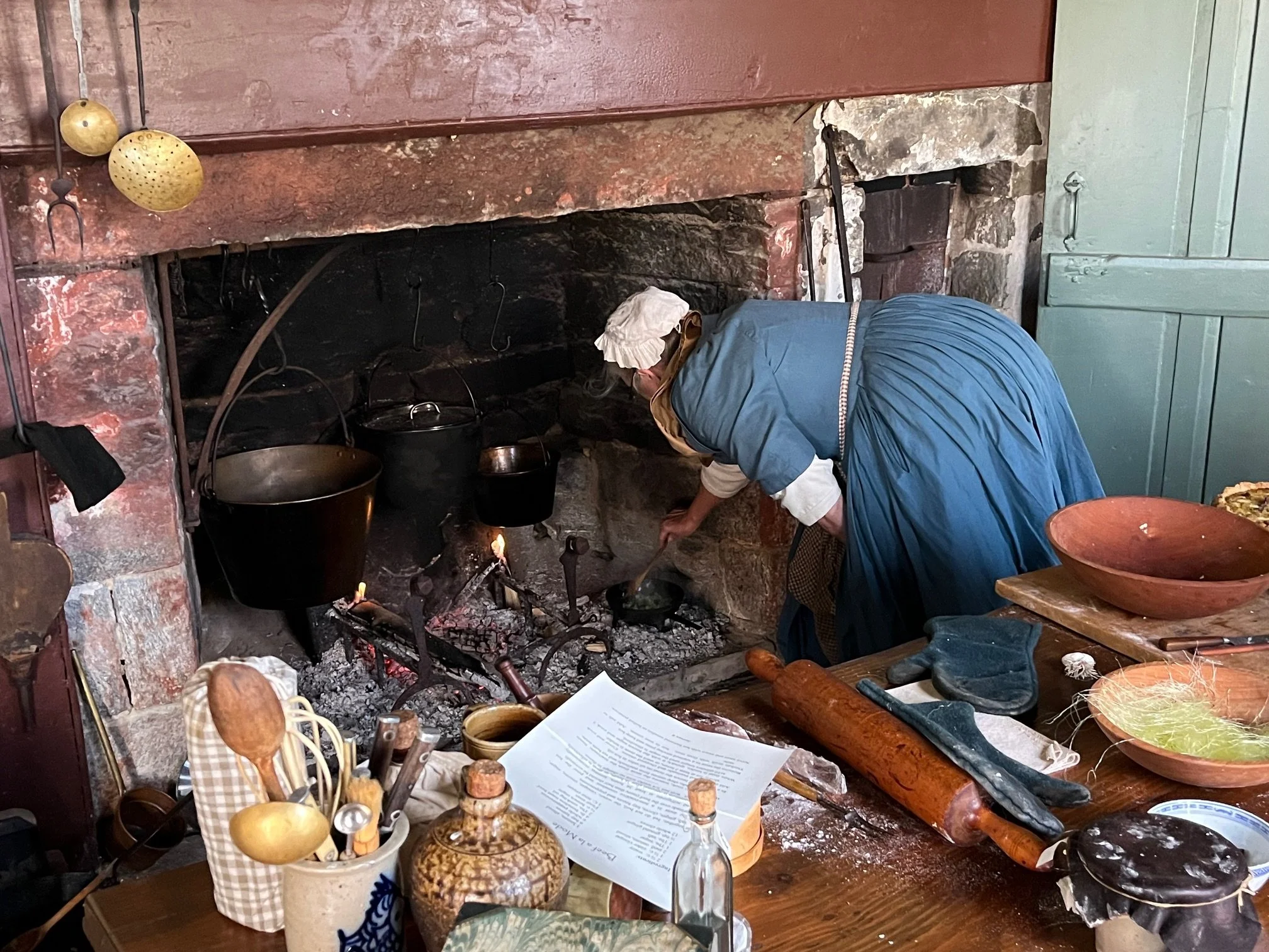 A woman dressed in historical clothing cooking over an open fire in a rustic kitchen with various utensils and dishes on the table.