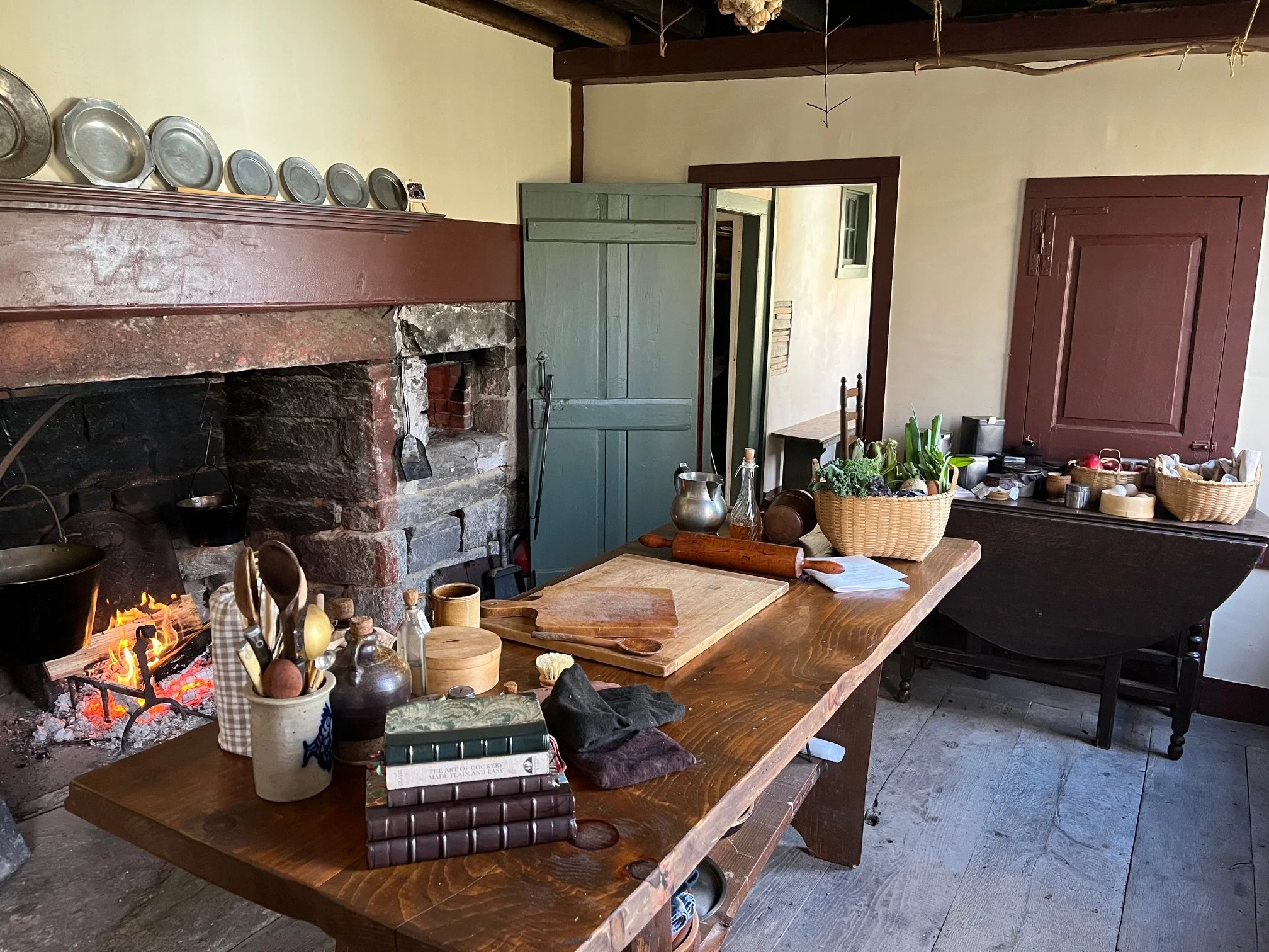 A rustic kitchen with a stone fireplace featuring a crackling fire and a large wooden table cluttered with cooking utensils, books, and a basket of vegetables. There is a green door, a closed wooden cabinet, and a side table with various kitchen items.