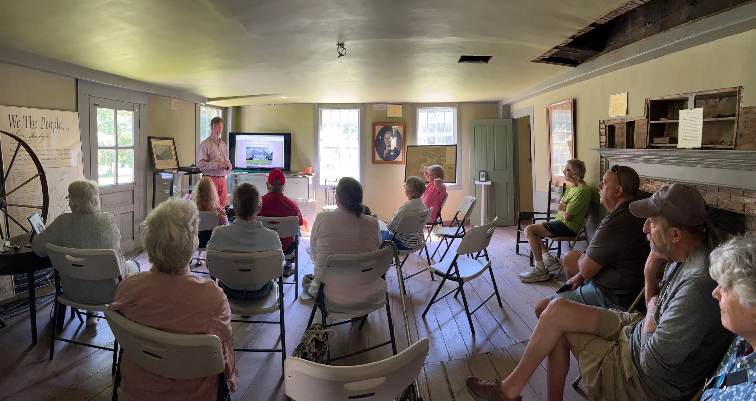 A group of people attending a presentation in a cozy, rustic room with yellow walls, wooden floors, and a brick fireplace. The presenter stands by a large screen displaying images, and the audience is seated on white folding chairs facing the presenter.