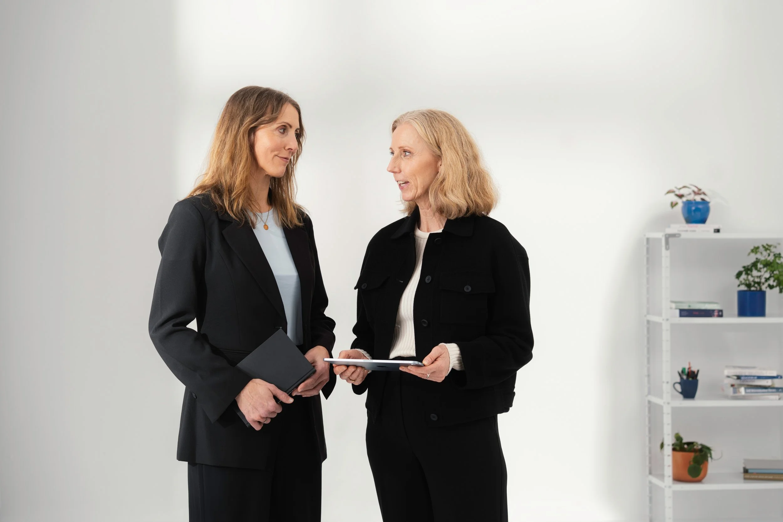 Two women in business attire having a conversation in an office with white walls and a white bookshelf with plants and books.
