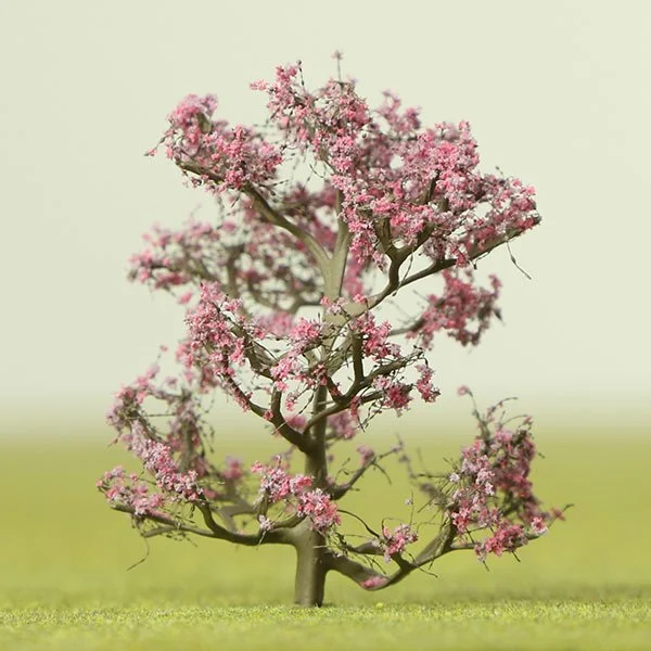 A small pink flowering model tree on a grassy field with a blurry sky background.