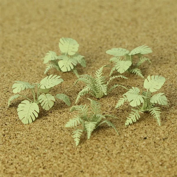 Artificial model plants resembling monstera leaves and fern leaves on a sandy surface.