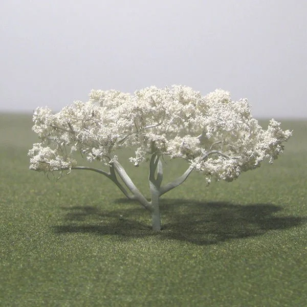 A model, white-blossomed tree on a grassy field with a plain sky background.