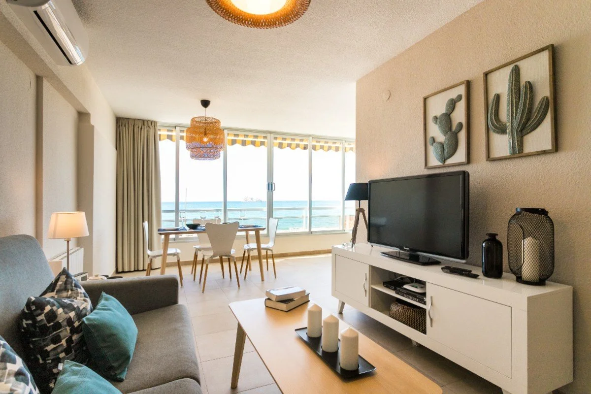 Sala de estar con televisor, sofá con cojines, mesa de centro con velas, comedor junto a vista al mar, decoraciones de cactus en la pared y cortinas.