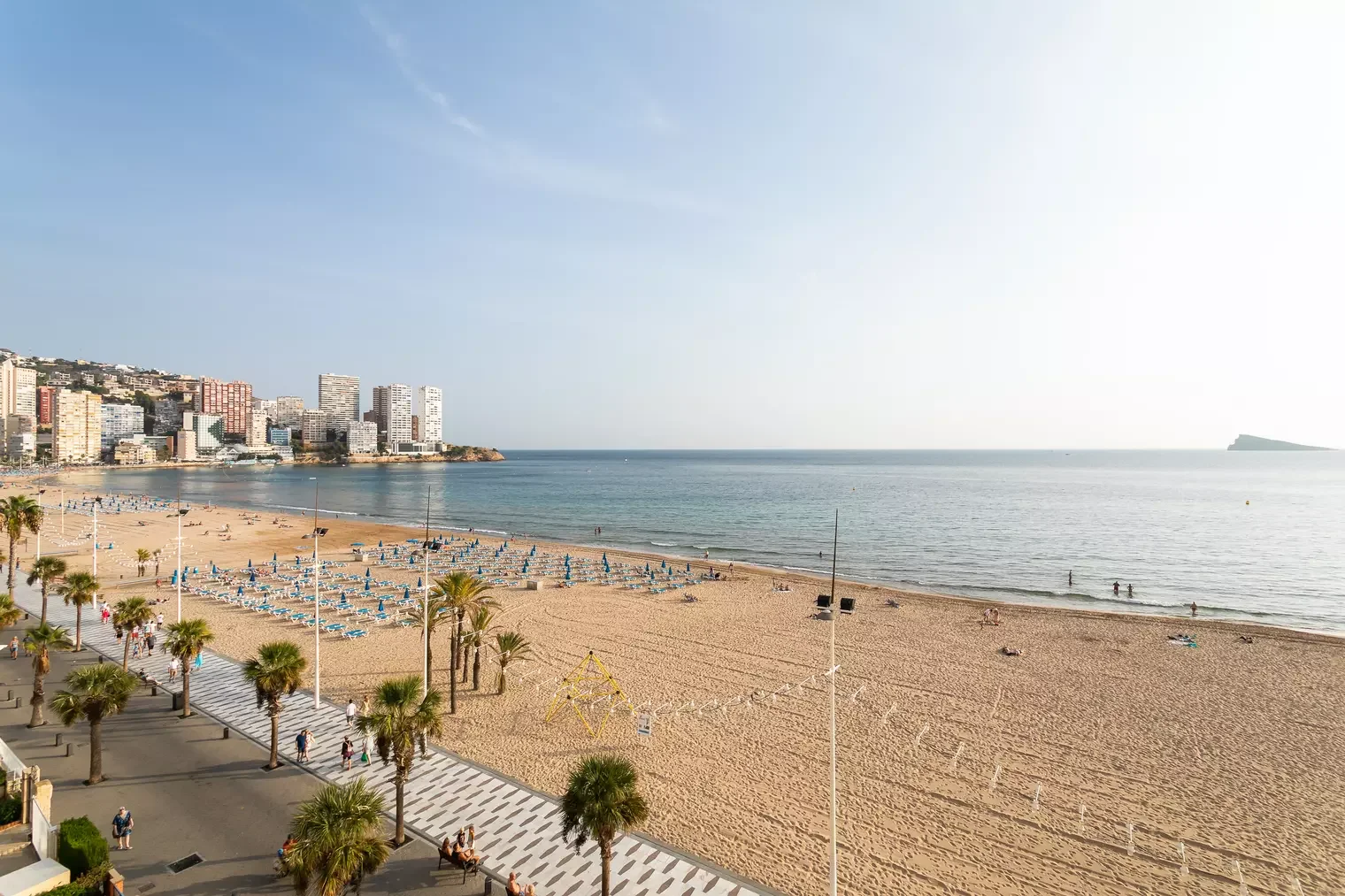 Vista de una playa urbana con arena dorada, palmeras y sombrillas azules, con edificios altos en el fondo y el mar tranquilo bajo un cielo despejado.