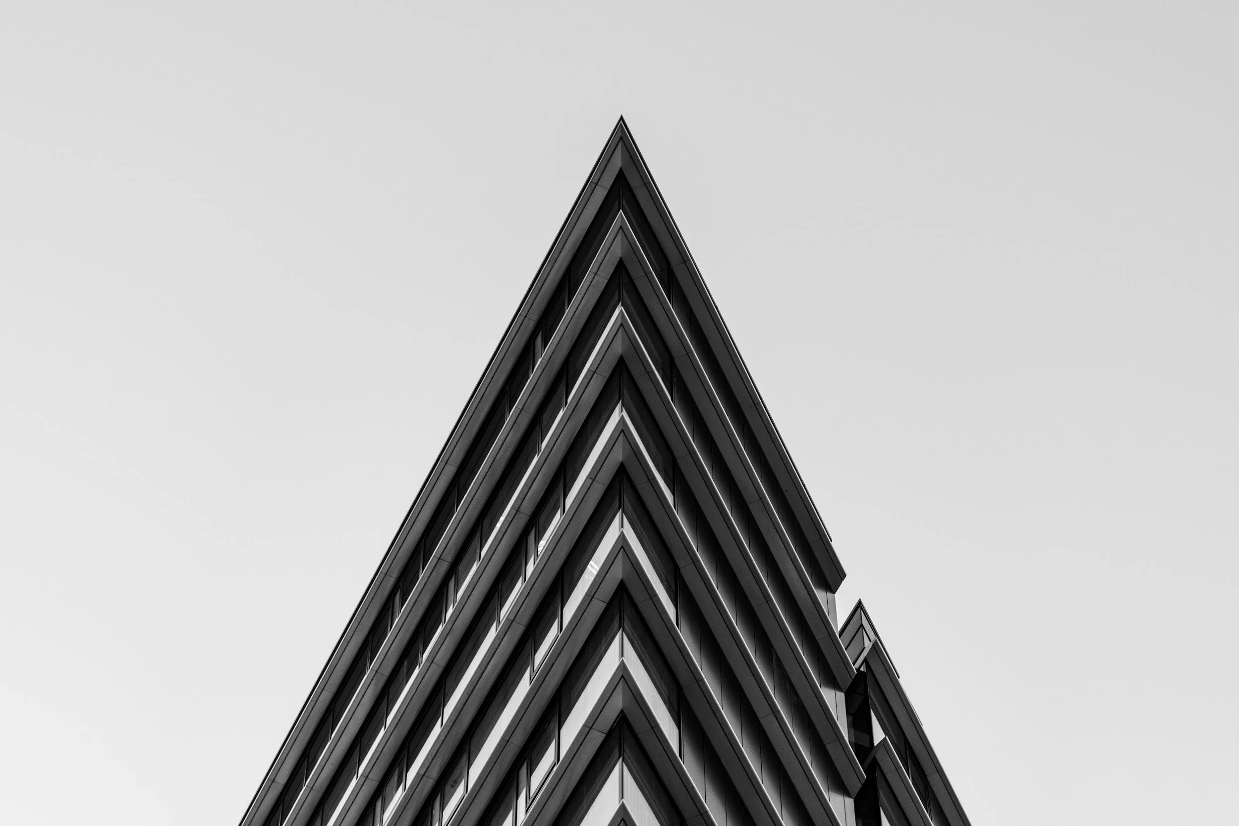 Black and white photograph of a modern building with sharp, angular lines, taken from below, showing the pointed corner of the structure against a clear sky.