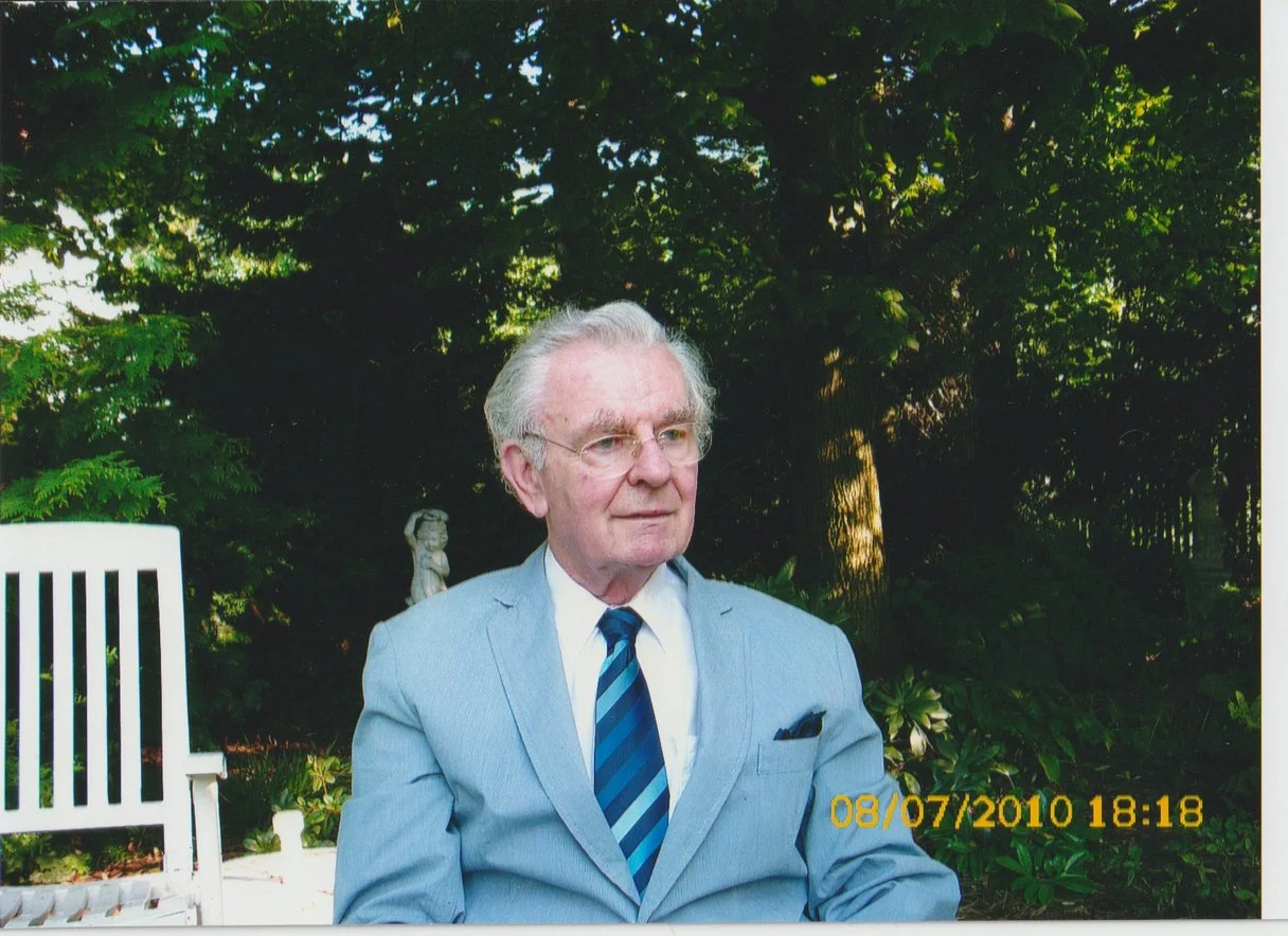 Älterer Mann mit weißen Haaren, Brille und Anzug sitzt im Garten vor Bäumen und Sträuchern, mit einer weißen Gartenbank im Hintergrund, Datum und Uhrzeit 09.07.2010 18:18