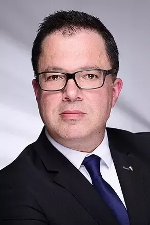 Porträt eines Mannes in formeller Kleidung mit Brille vor neutralem Hintergrund.