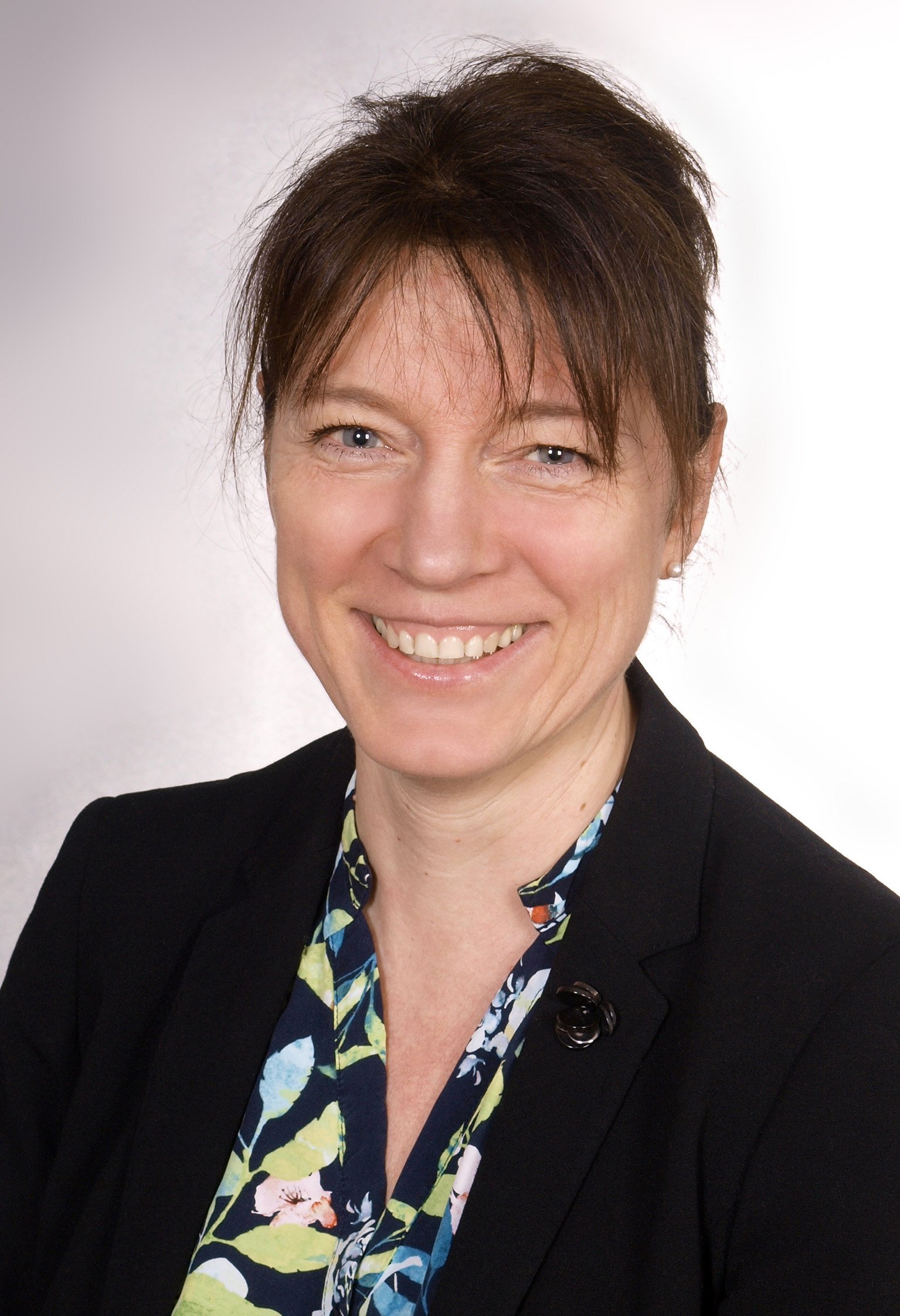 Porträt einer Frau mit braunem, kurzen Haar, blauen Augen und einem lächelnden Gesicht, trägt eine schwarze Jacke und ein blumiges Hemd.