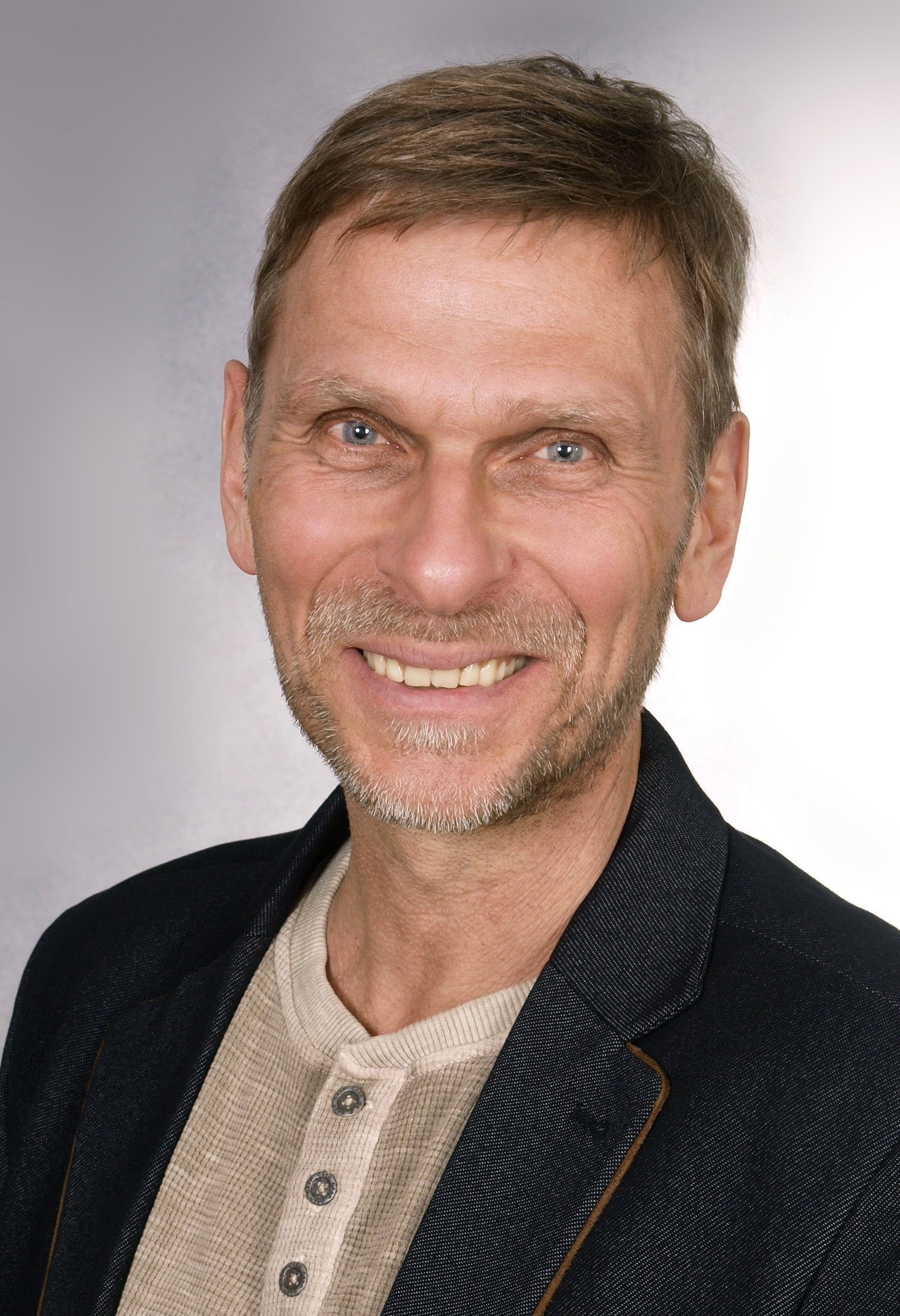 Porträt eines Lächelnden Mannes mit braunen Haaren, blauen Augen und Bart, im schwarzen Jackett und hellen Hemd, vor hellem Hintergrund.