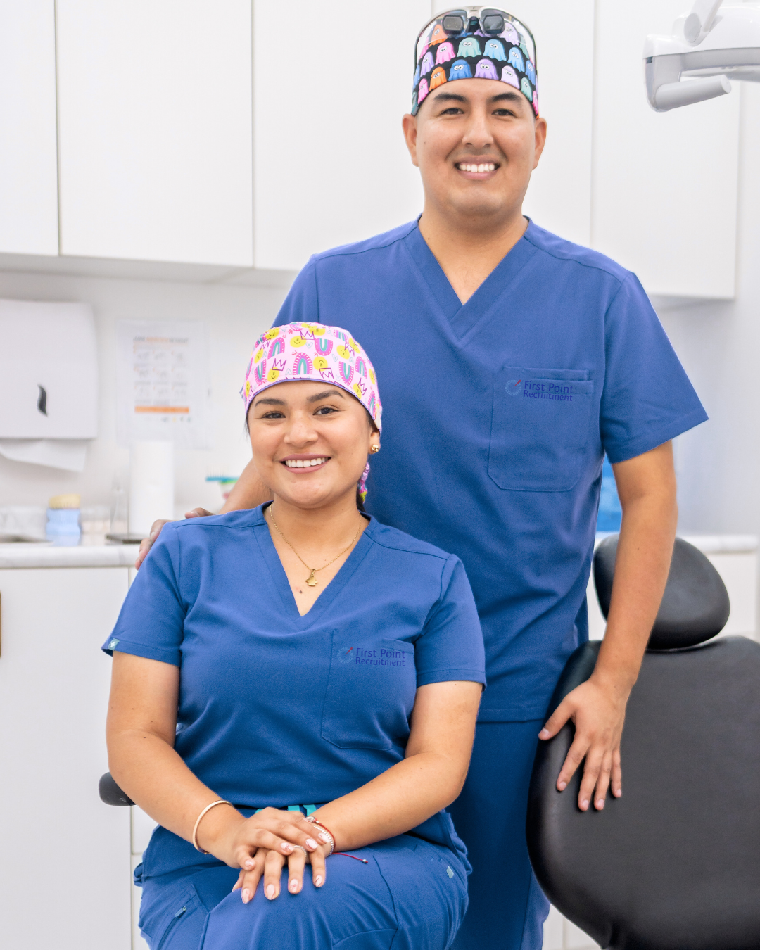 Two smiling healthcare professionals in blue scrubs, a woman seated and a man standing, in a medical setting.