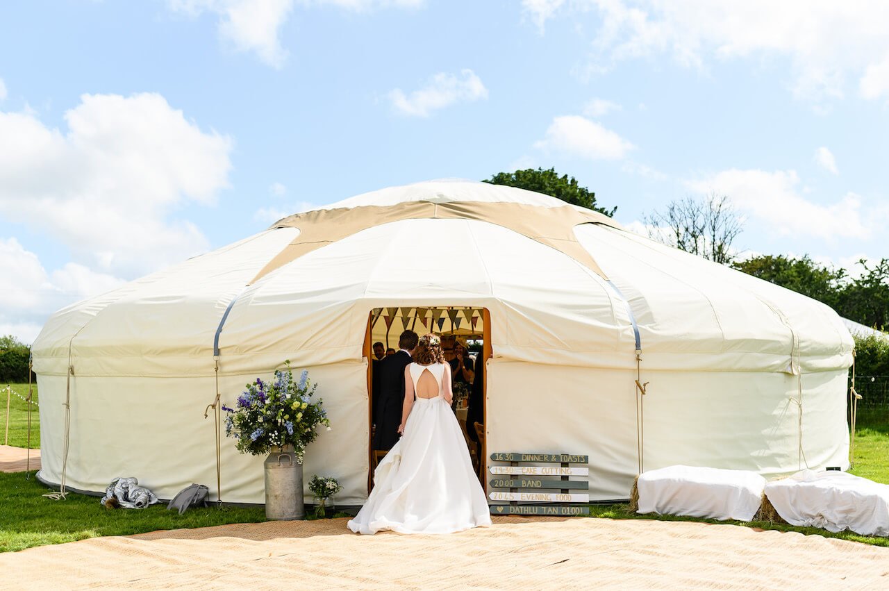 A bride in a white wedding dress walking into a large outdoor event tent on a sunny day, with a group of people inside and decorative flower arrangements outside.