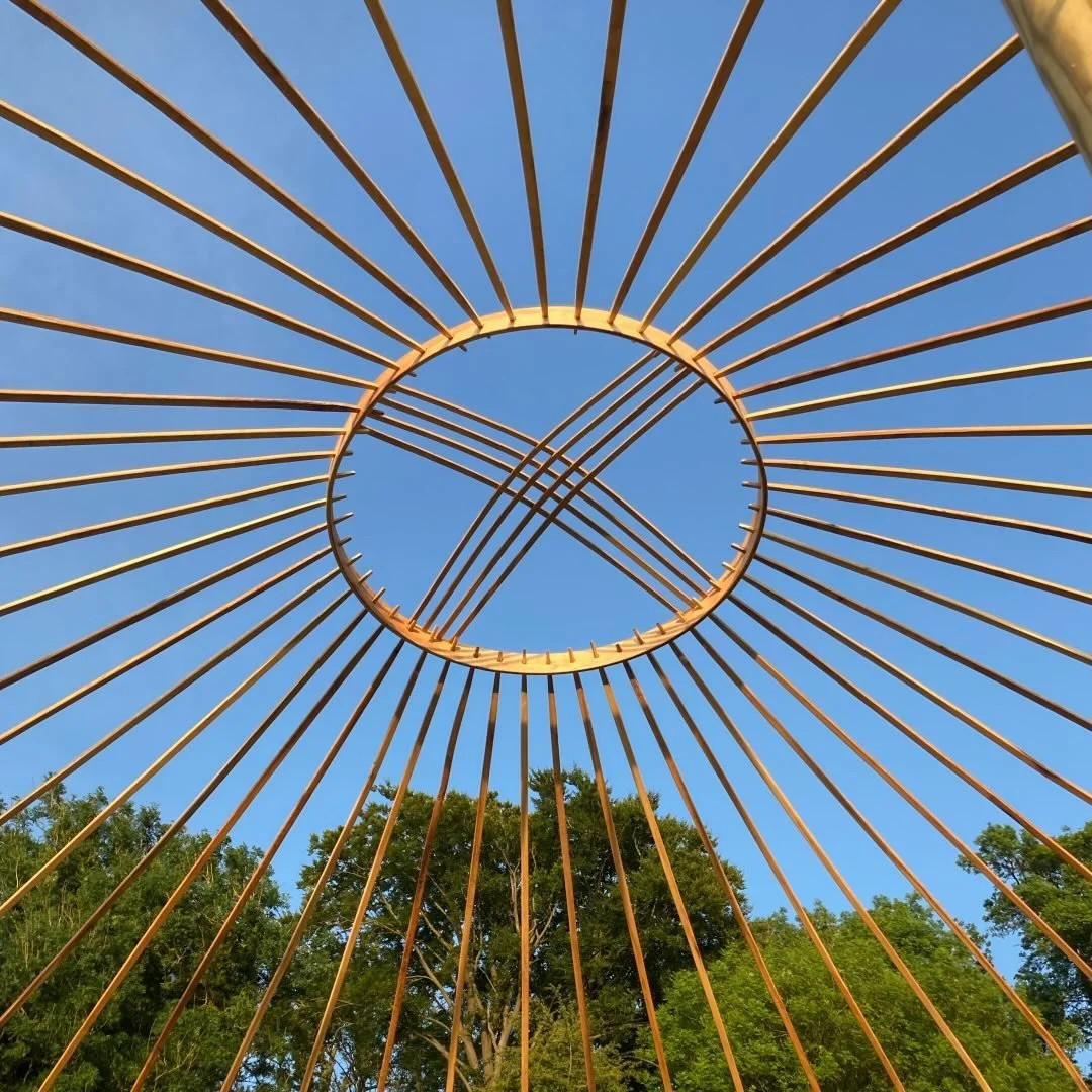 View from below of a circular metal sculpture with radiating spokes against a clear blue sky. Green trees are visible at the bottom of the image.