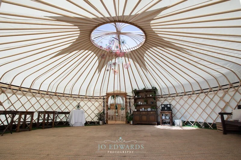 Interior of a large, circular, canvas yurt with wooden lattice walls, decorated with hanging pink flowers, potted plants, a wooden sideboard, and a small table, beneath a sky-visible circular skylight.