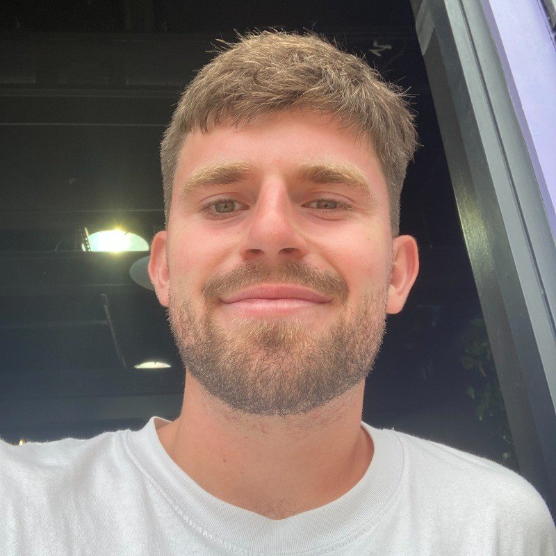 A young man with short light brown hair and a full beard is smiling at the camera. He is wearing a white T-shirt and standing in front of a dark background with some reflective surfaces.