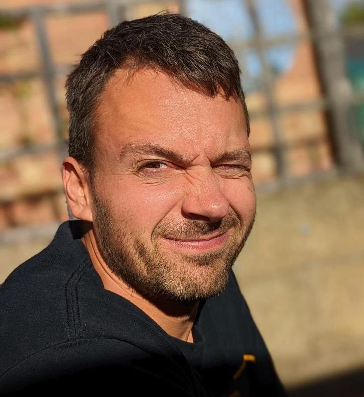 A man with short dark hair and a beard squinting and smiling at the camera outdoors in sunlight.