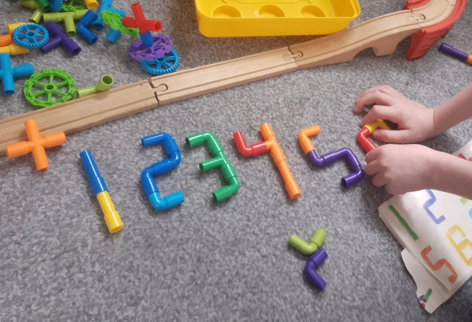 Colorful plastic numbered shapes arranged to form the sequence 1, 2, 3, 4, 5, 6 on a gray carpet, with a child's hands assembling a purple number 4.