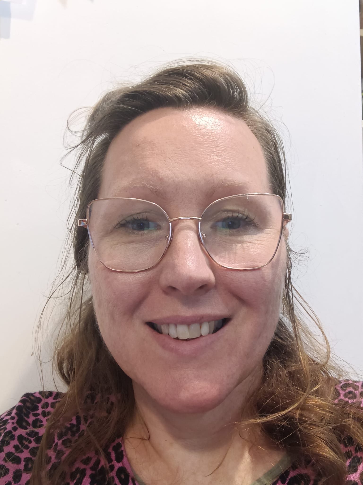 A woman with wavy brown hair, wearing clear glasses and a pink leopard print top, smiling at the camera against a white background.