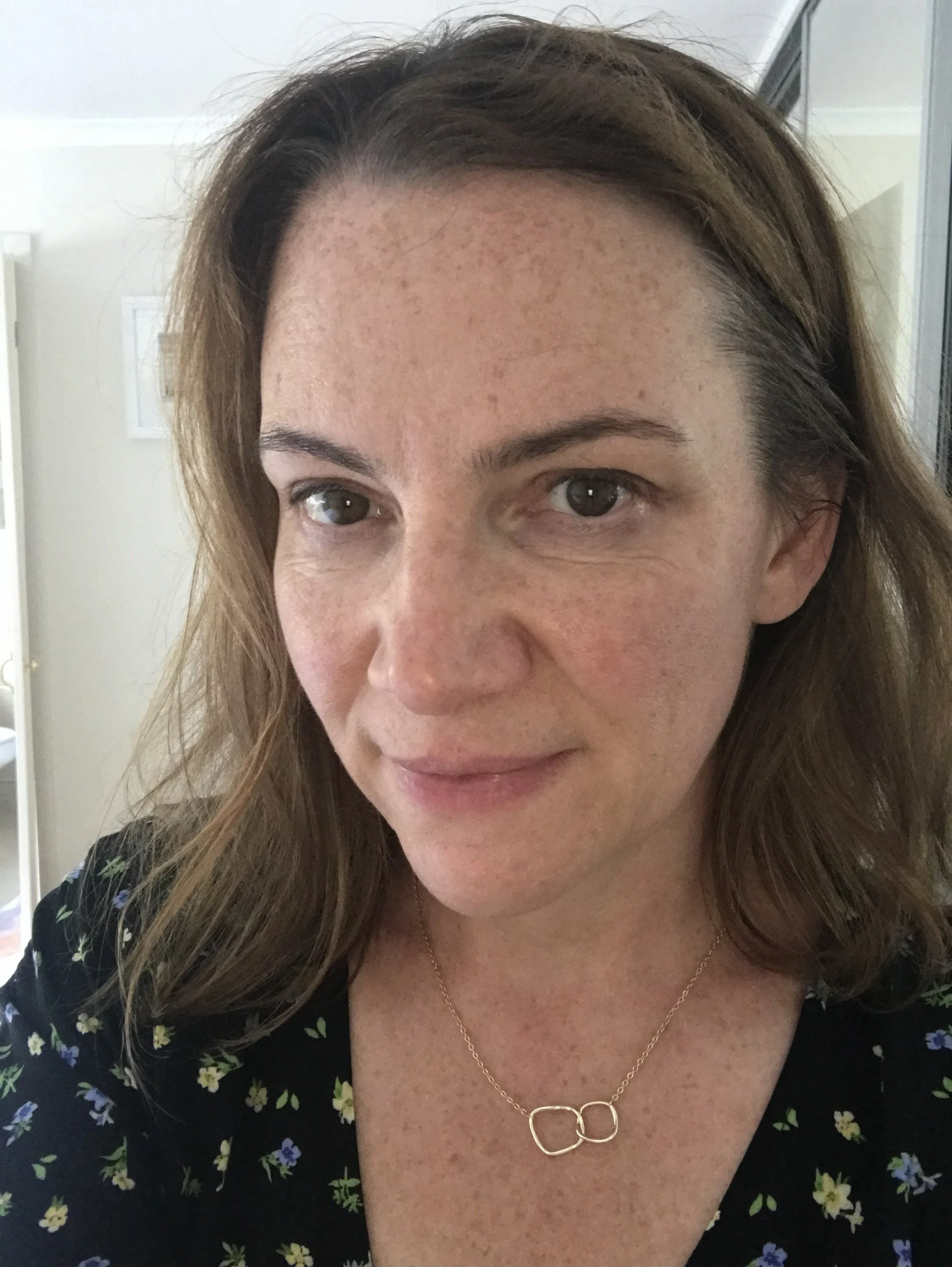 Close-up of a woman with light skin, freckled face, brown hair, wearing a gold necklace with two interconnected circles, and a black top with a small floral pattern.