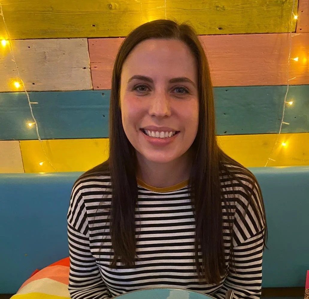 A smiling woman with long brown hair, wearing a black and white striped shirt, sitting in front of a colorful wooden wall with pastel blocks and string lights.