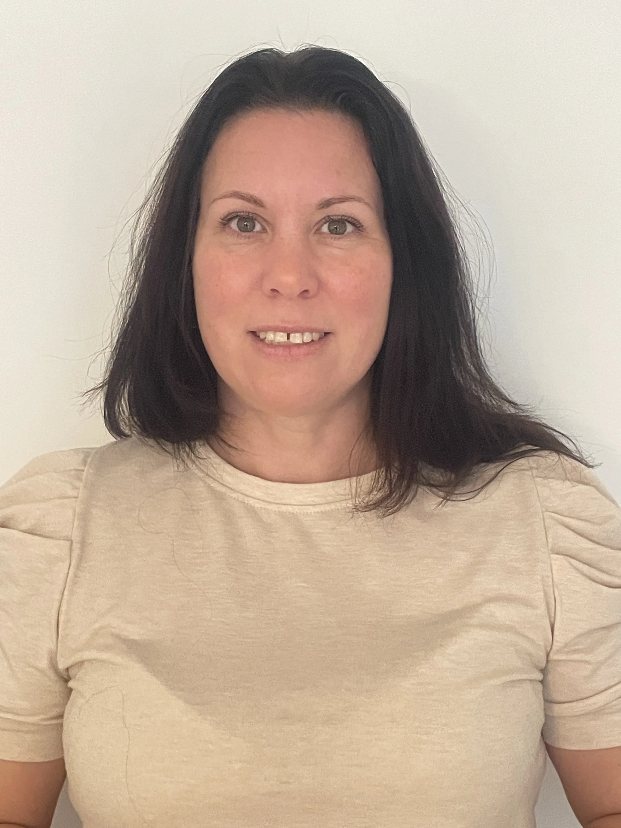 A woman with shoulder-length dark hair, light skin, wearing a beige top with puffed sleeves, standing against a plain white background.