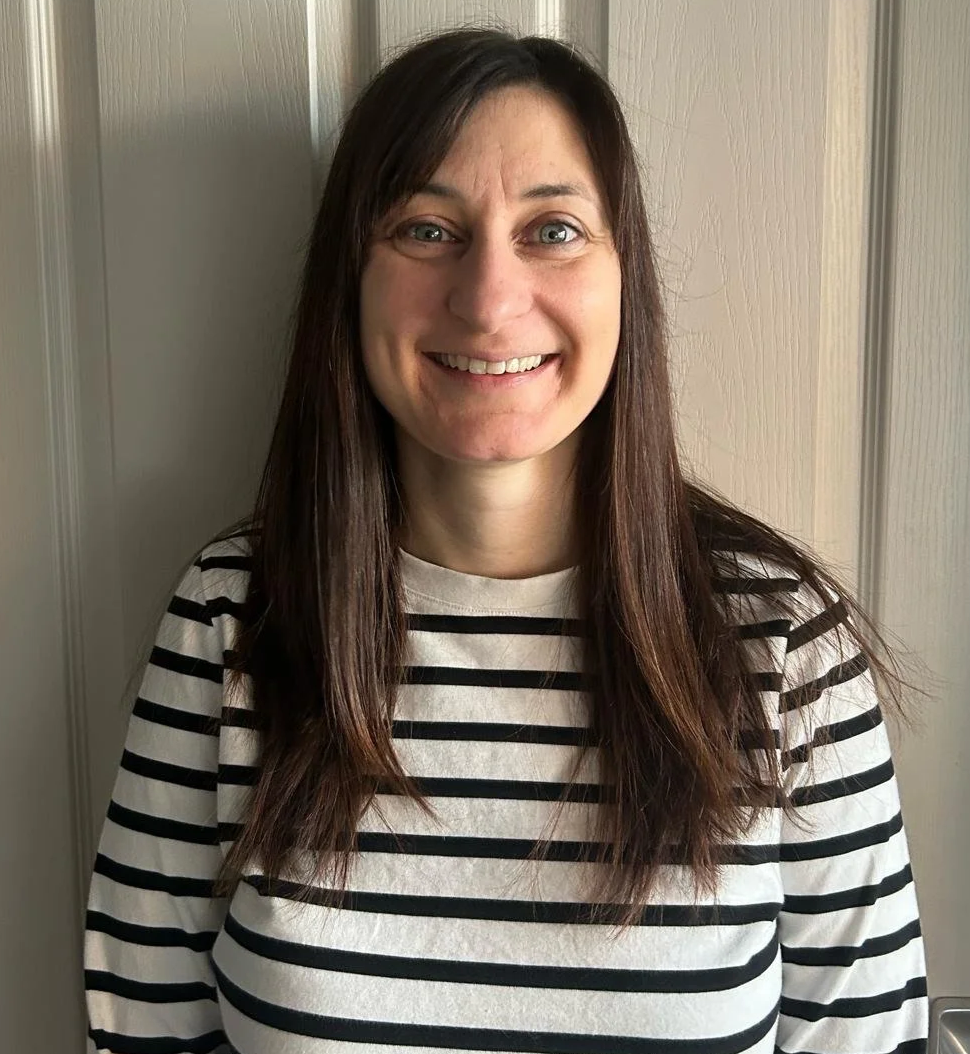 A woman with long brown hair wearing a white shirt with black horizontal stripes, smiling in front of a beige wall.