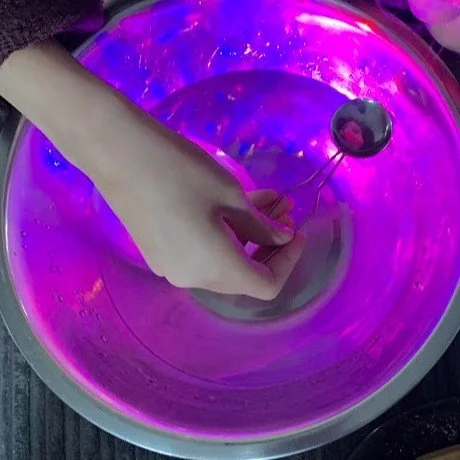 A hand holding a metal spoon with a reflective ball at the end, stirring a glowing, purple-lit bowl of liquid.
