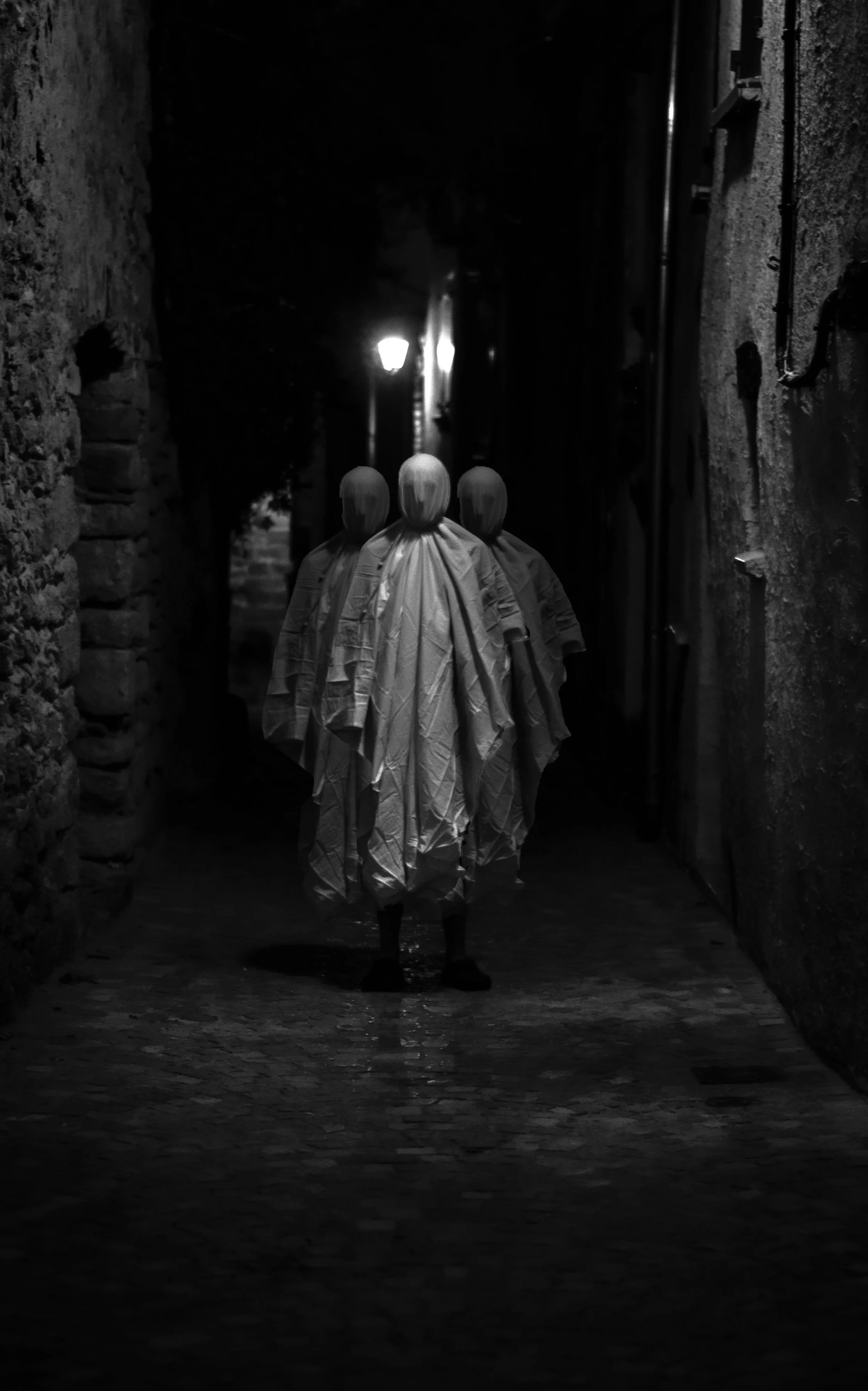Three mannequins dressed in robes walking through a dark alley at night, with a street lamp illuminating the background.