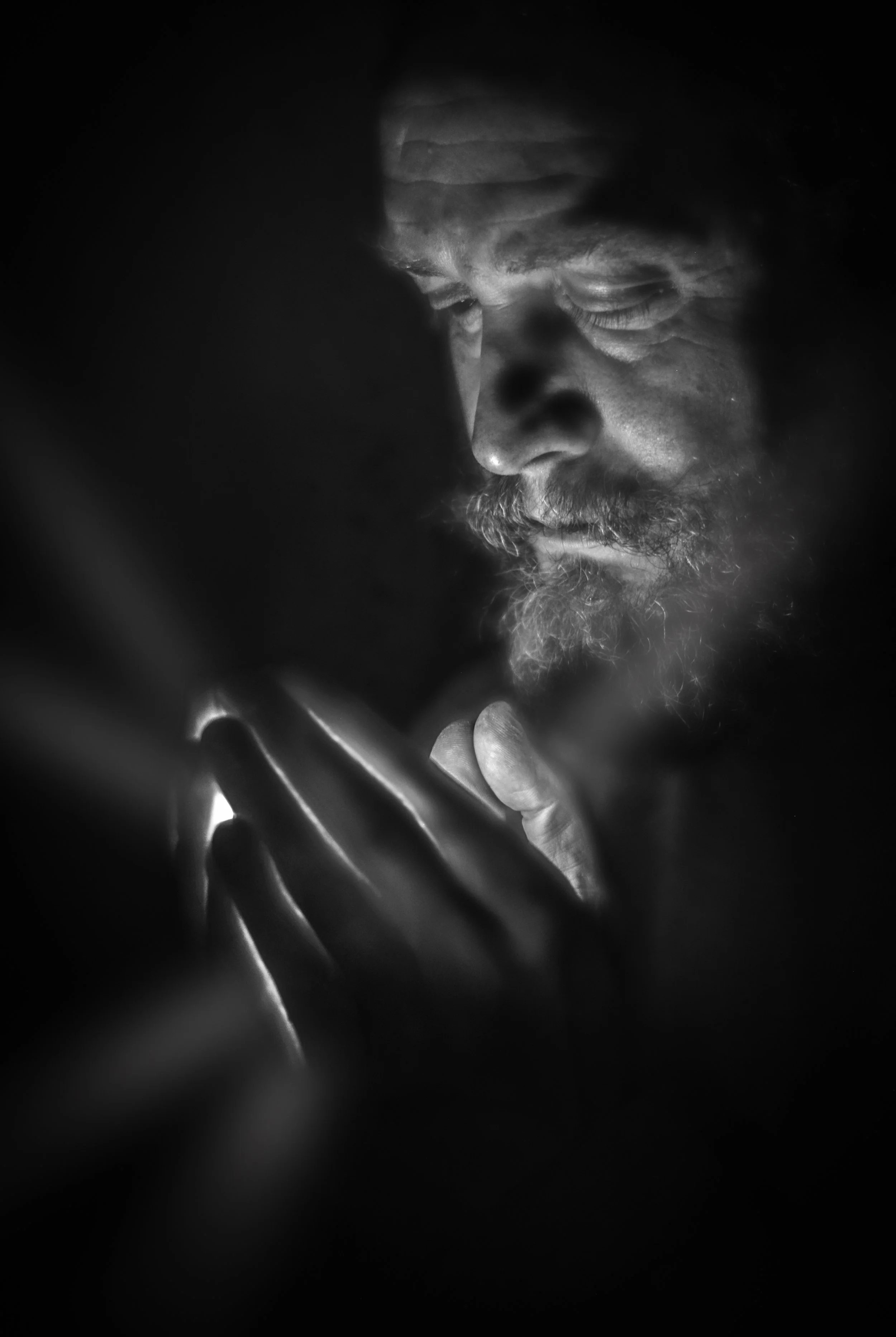 Black and white close-up of a bearded man's face, with his eyes closed, holding his chin with his hand, illuminated softly in darkness.