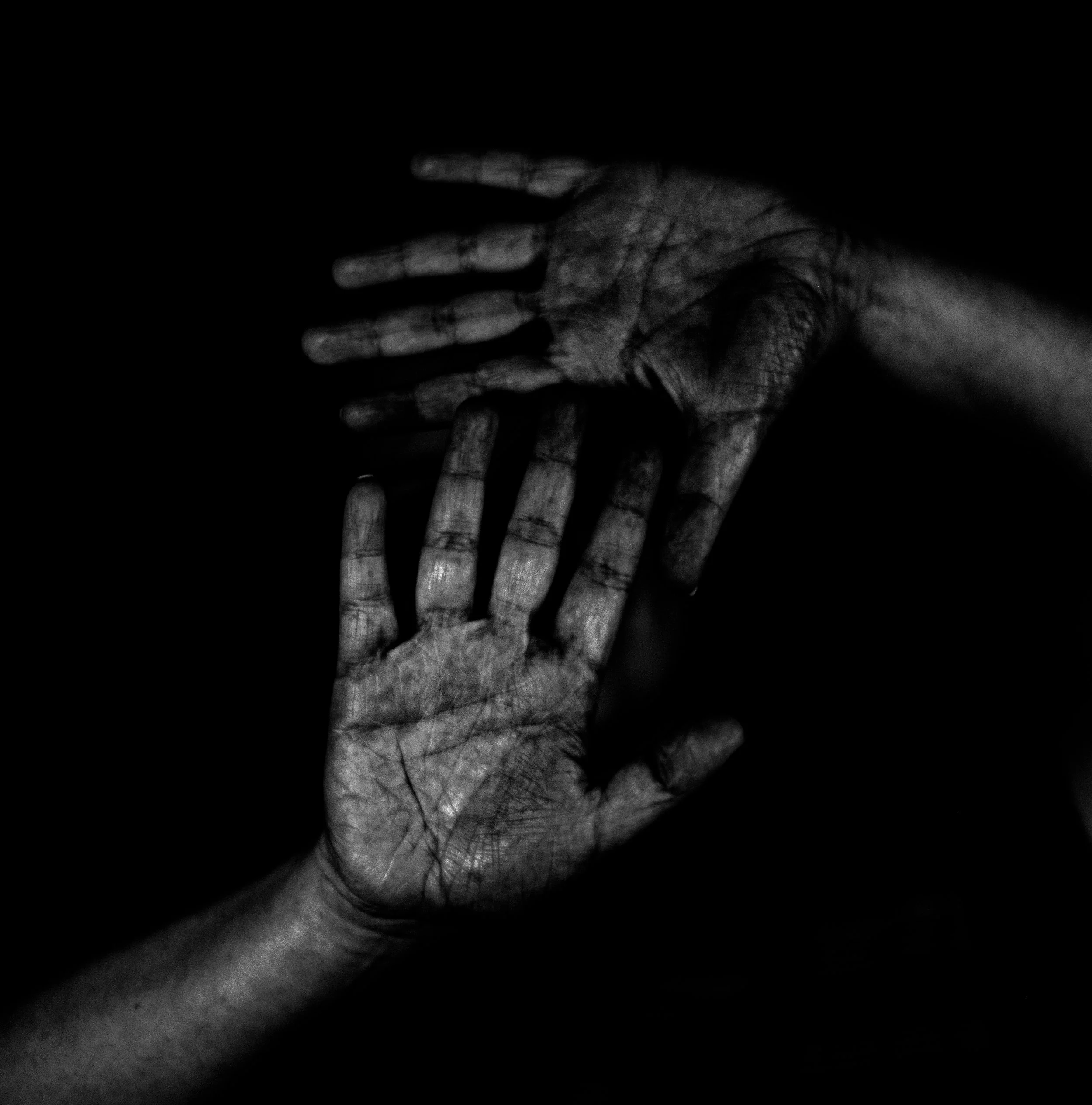 Black and white image of two hands, one stacked over the other, with visible lines and textures, set against a dark background.