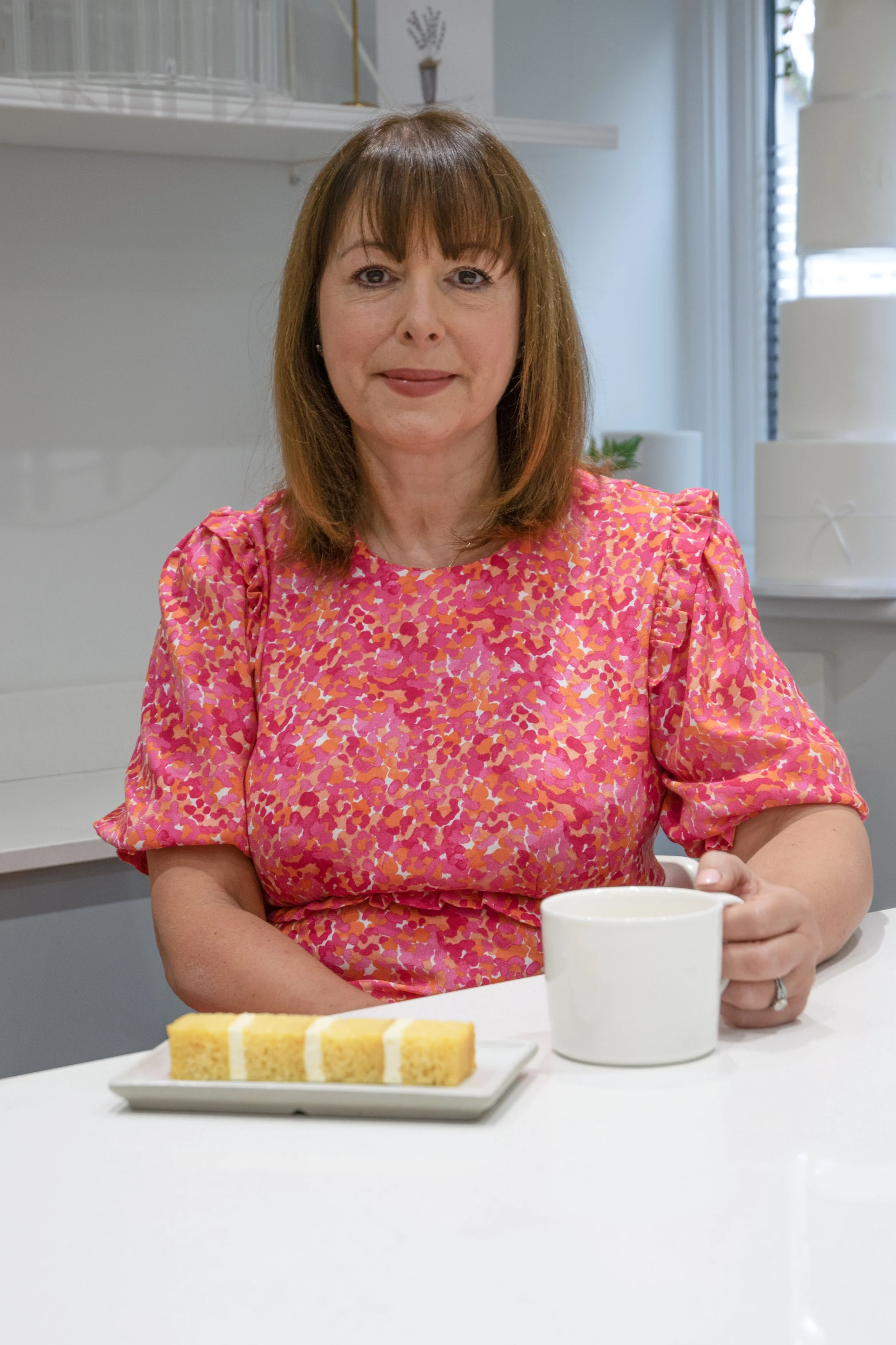 A woman with shoulder-length brown hair wearing a pink and orange patterned blouse, sitting at a white table with a white mug and a small plate of sliced yellow cake in front of her, in a bright modern kitchen.