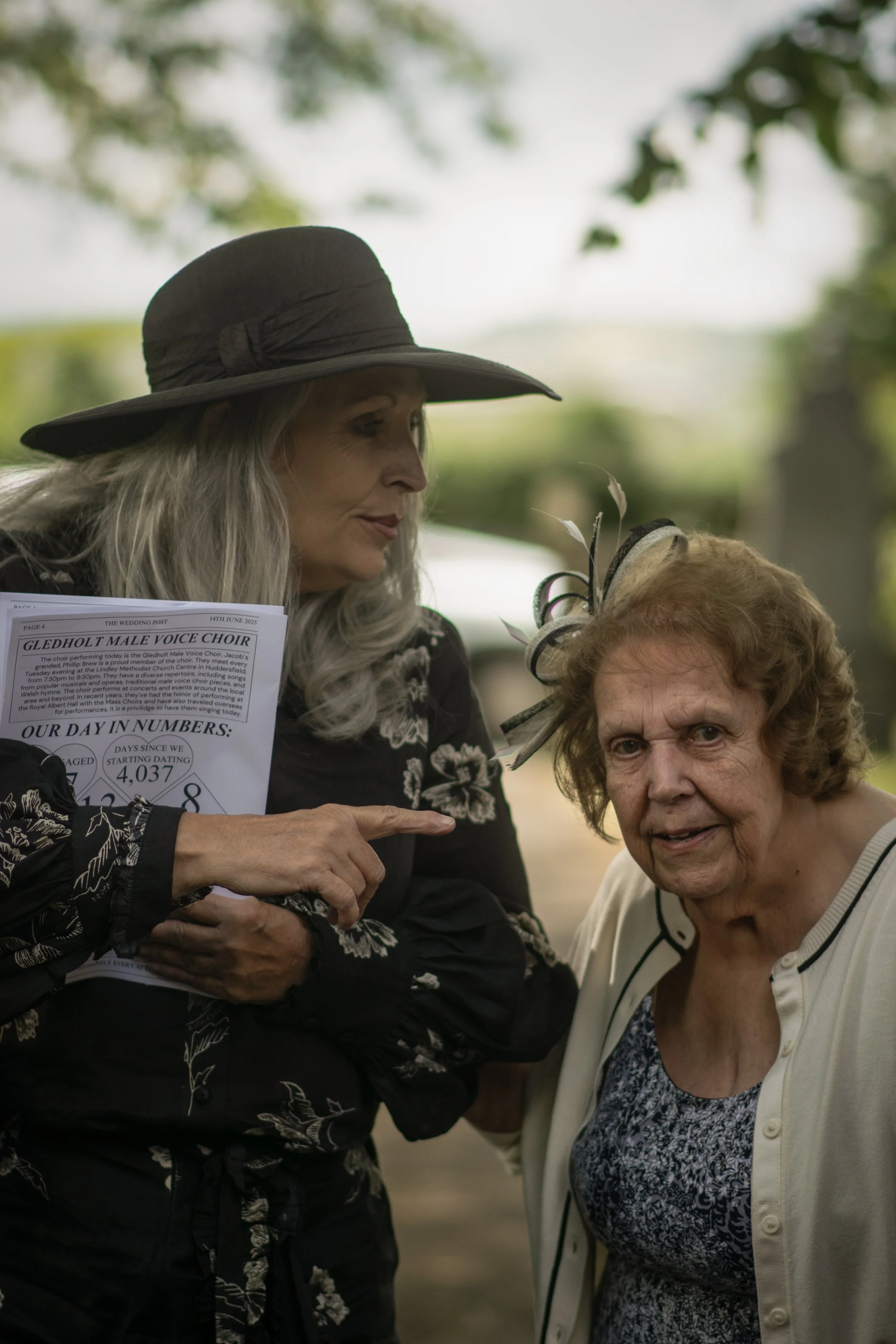 Two women having a conversation outdoors, one is wearing a black wide-brimmed hat and holding a paper, the other is wearing a fascinator hat and a light-colored cardigan.