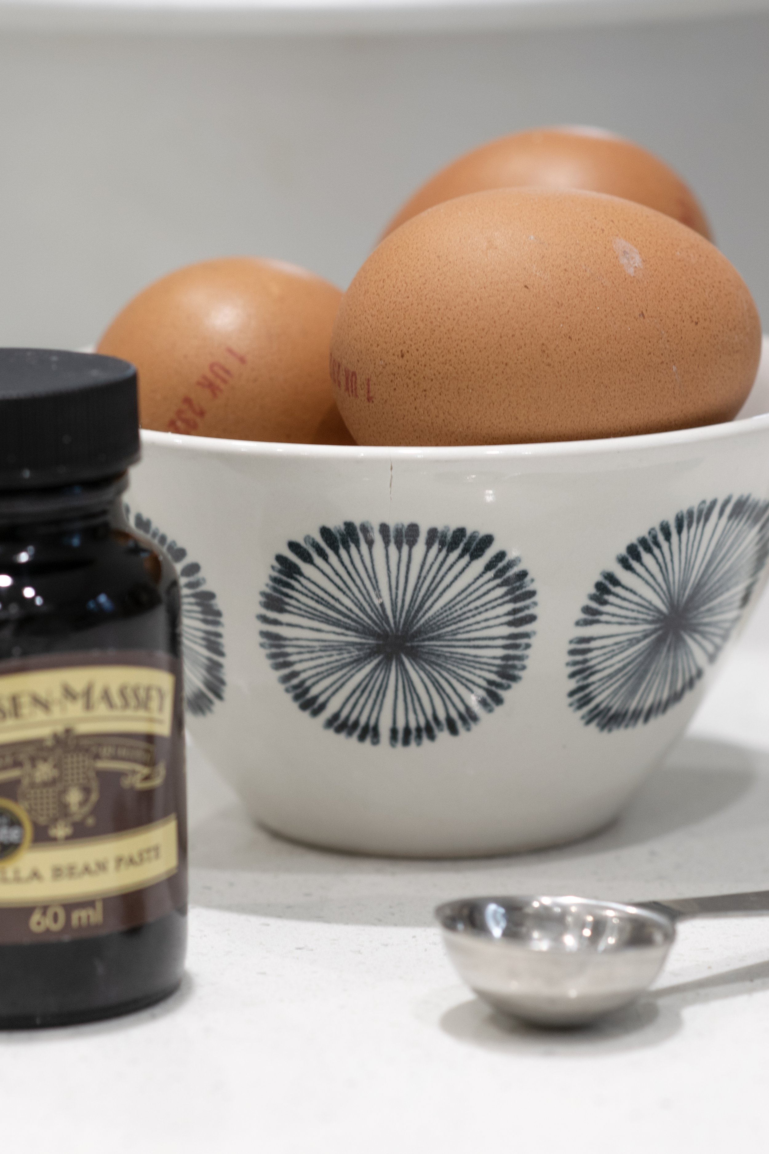 A white bowl with black firework patterns holding three brown eggs, a small brown bottle of Benemassey vanilla bean paste, and a metal measuring spoon on a white surface.