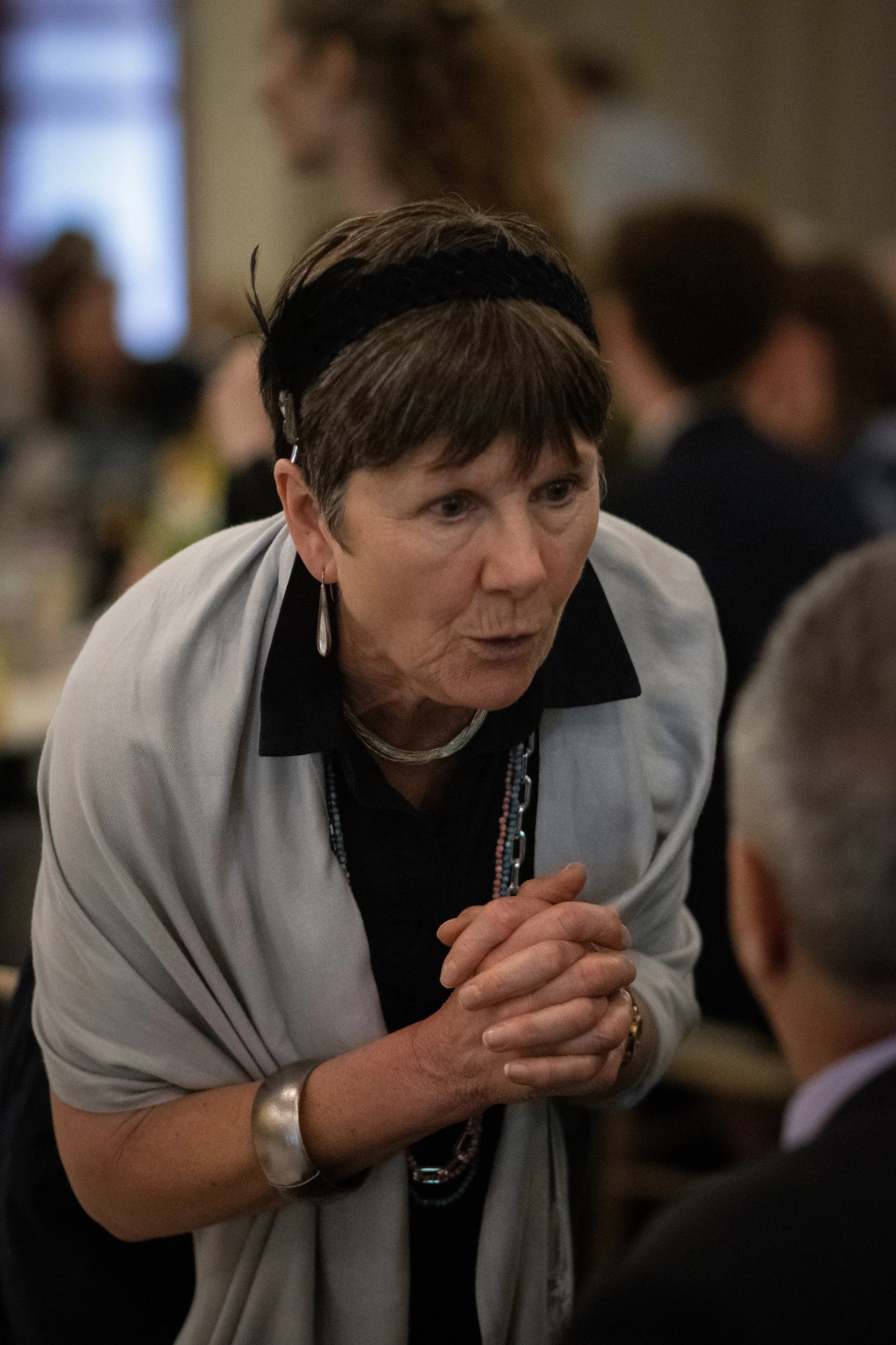 A woman with short brown hair, wearing a light gray jacket, black shirt, and jewelry, is leaning forward and speaking intently to a man. She has her hands clasped together and appears to be engaged in a serious conversation at an indoor event with other blurred attendees in the background.