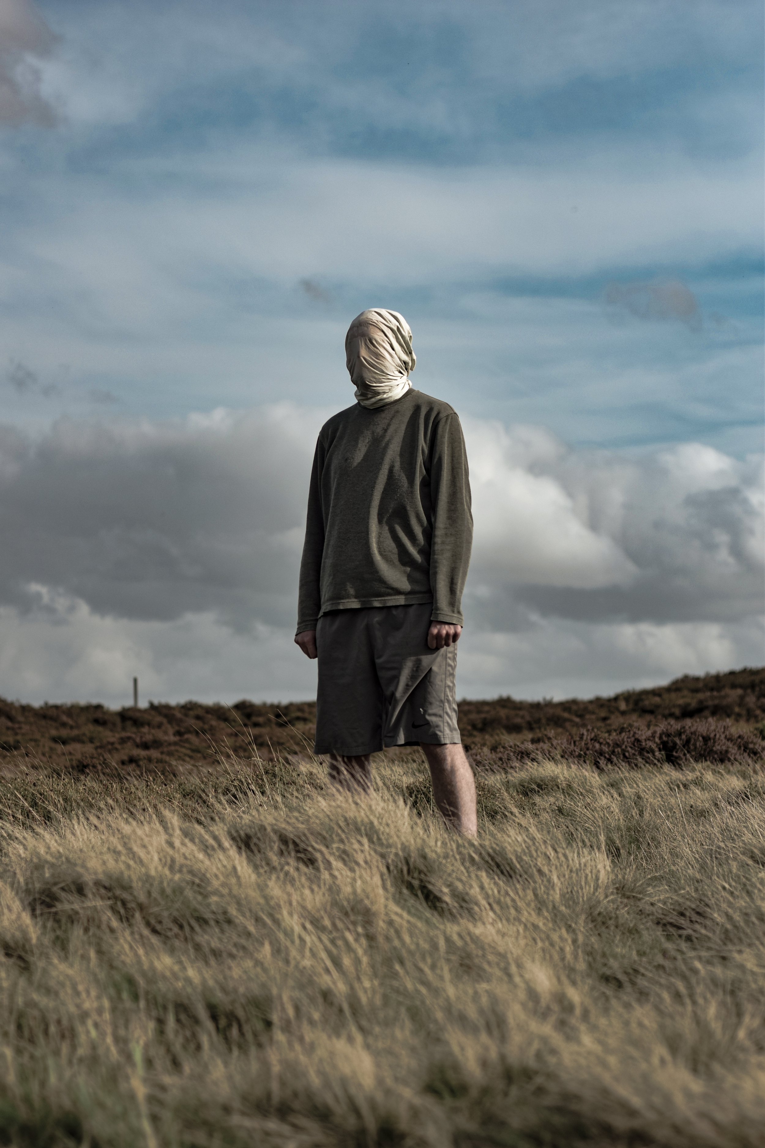 A person dressed in a hoodie and shorts with their head wrapped in a cloth, standing in a grassy field under a cloudy sky.