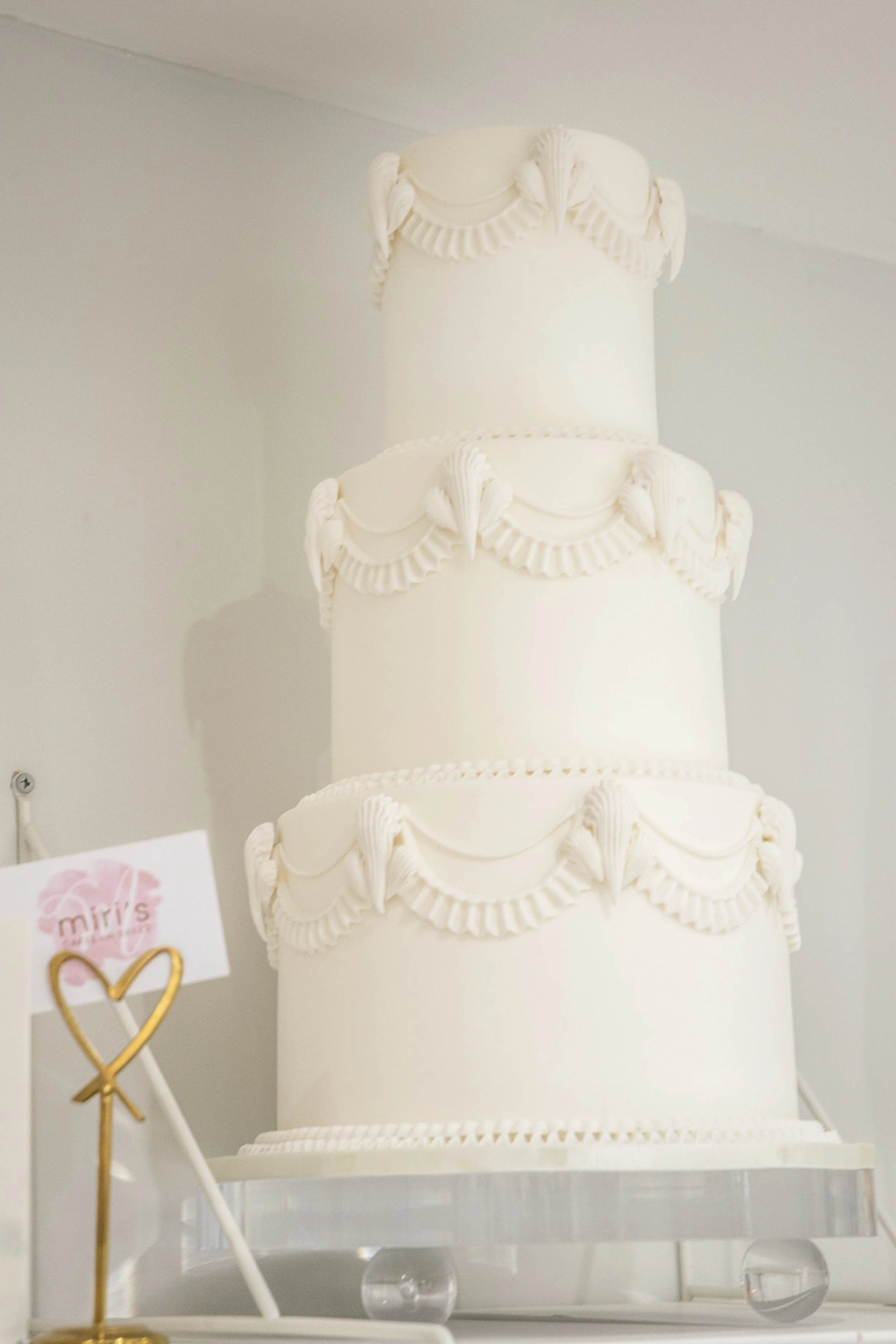 Three-tiered white wedding cake decorated with white frosting garlands and rosettes, displayed on a clear stand.