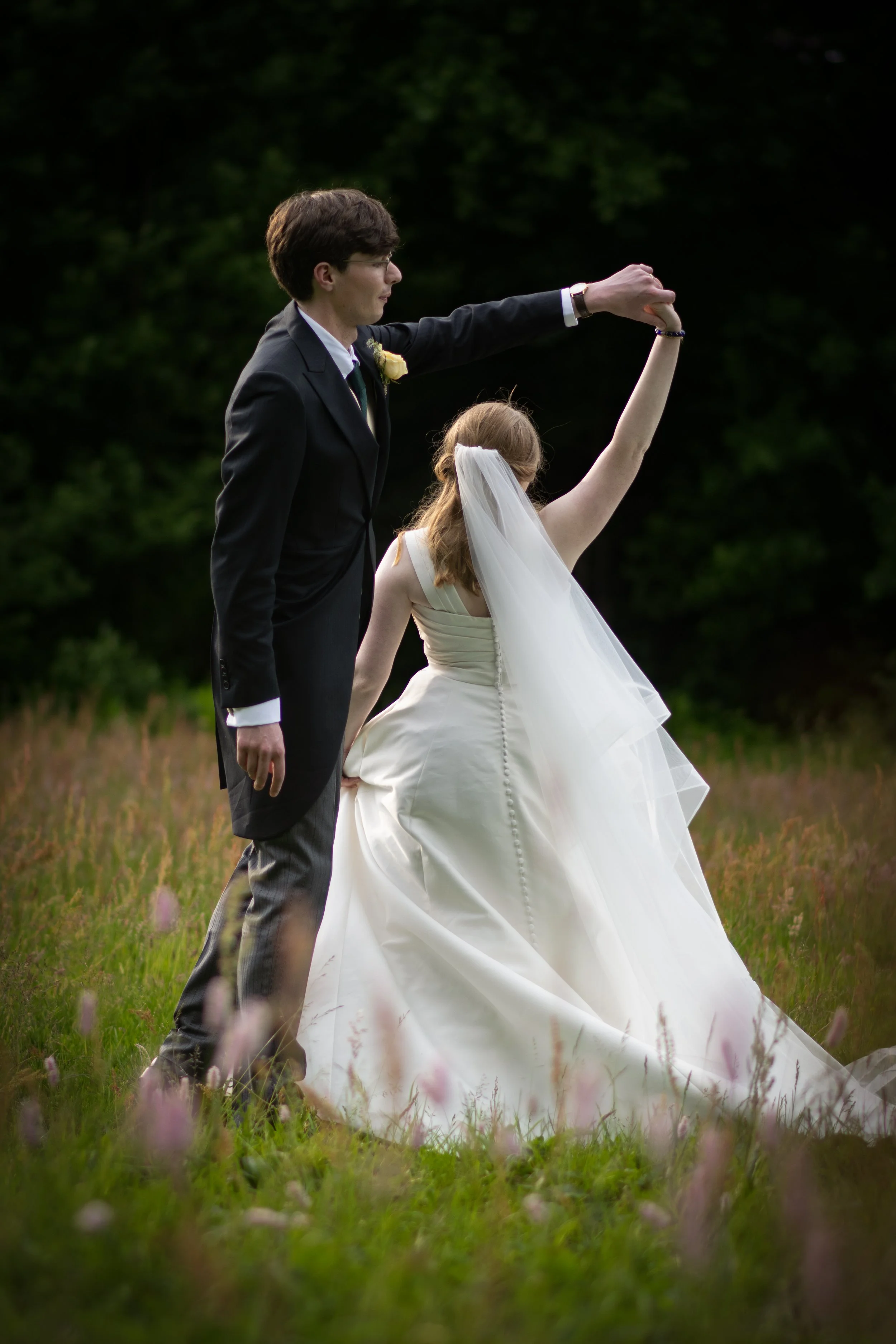 A bride and groom dancing outdoors in a grassy field during their wedding, with the groom holding the bride's hand as she spins.