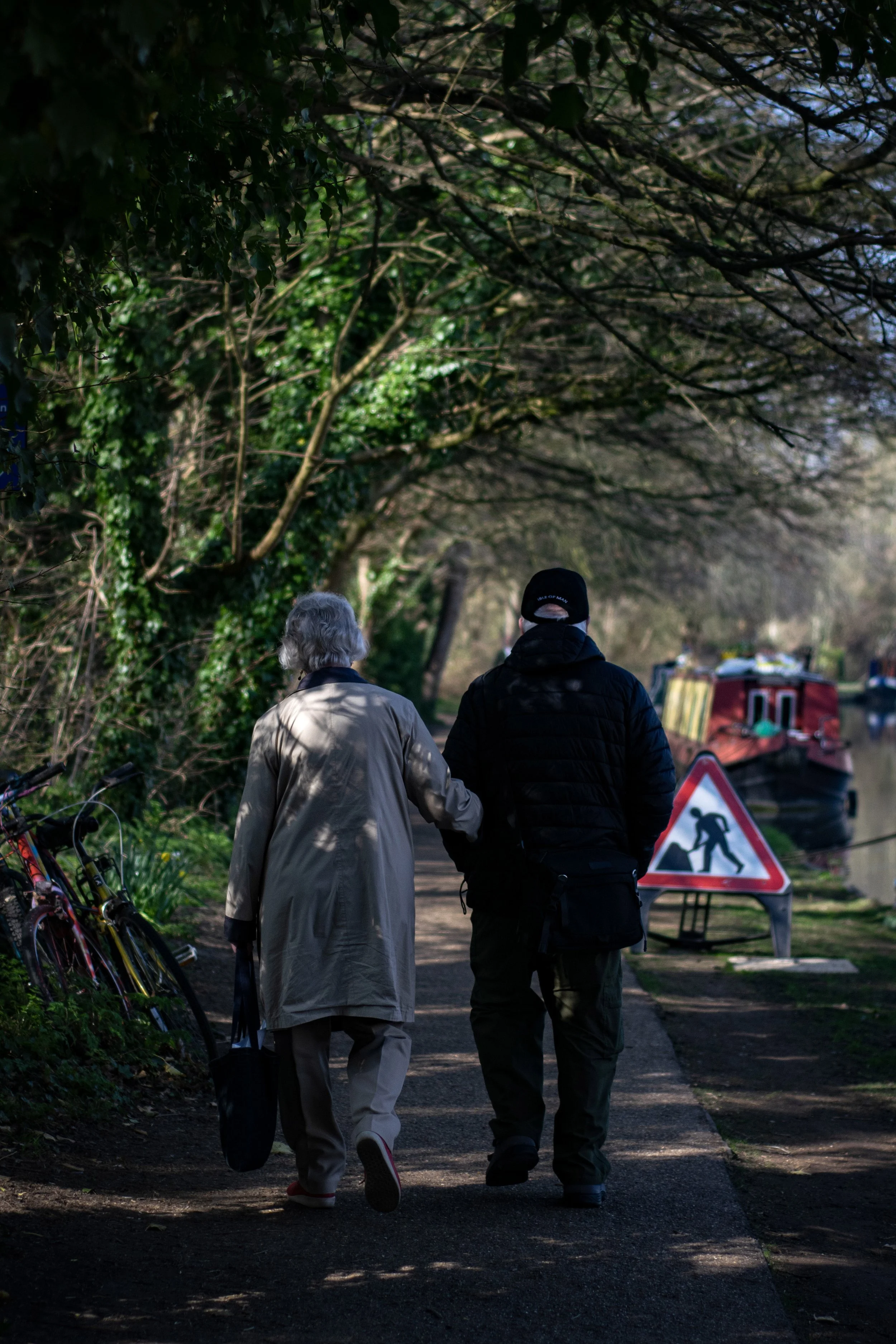 An elderly woman with gray hair and a man with dark hair and a cap walk together on a shaded riverside path. Bicycles are parked on the side, and a construction or work sign is visible near boats on the river.