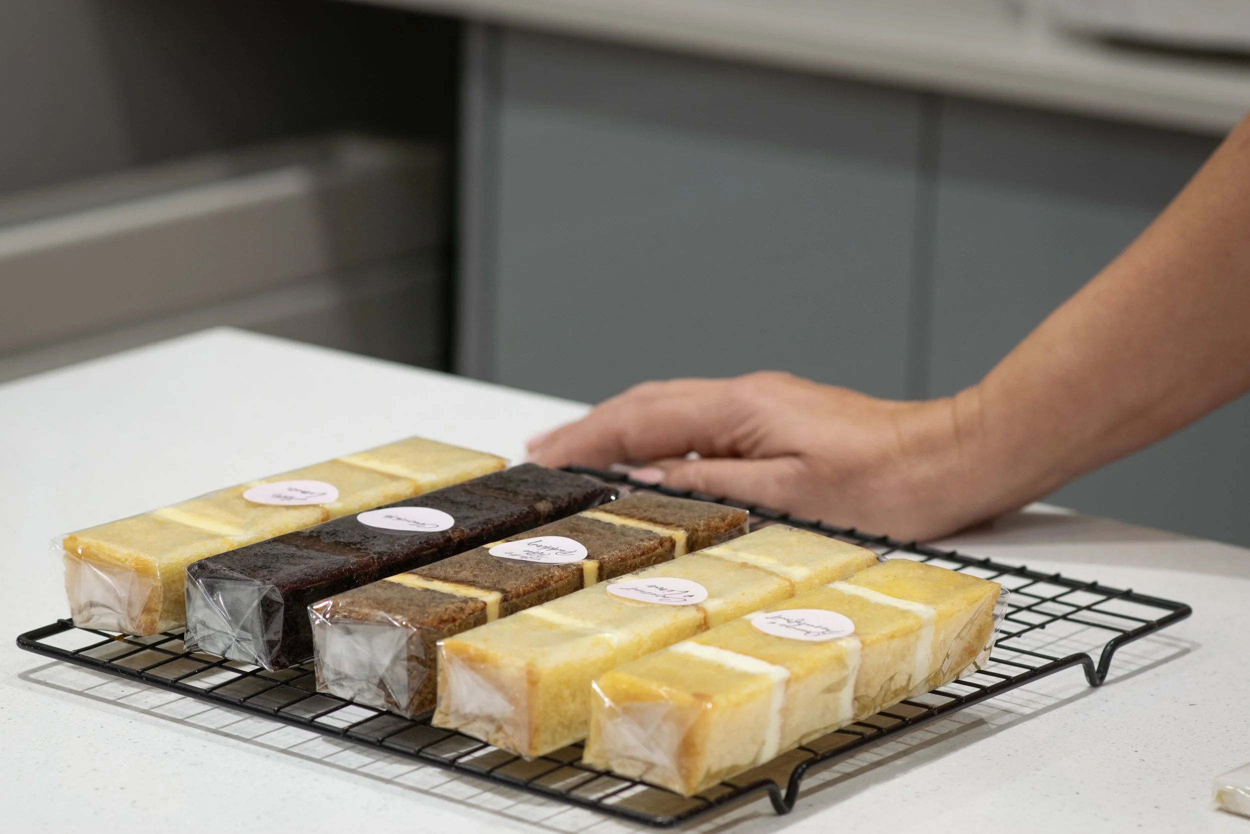 Assorted layered dessert bars wrapped in clear plastic on a cooling rack, with one person’s hand nearby on a white countertop in a kitchen setting.