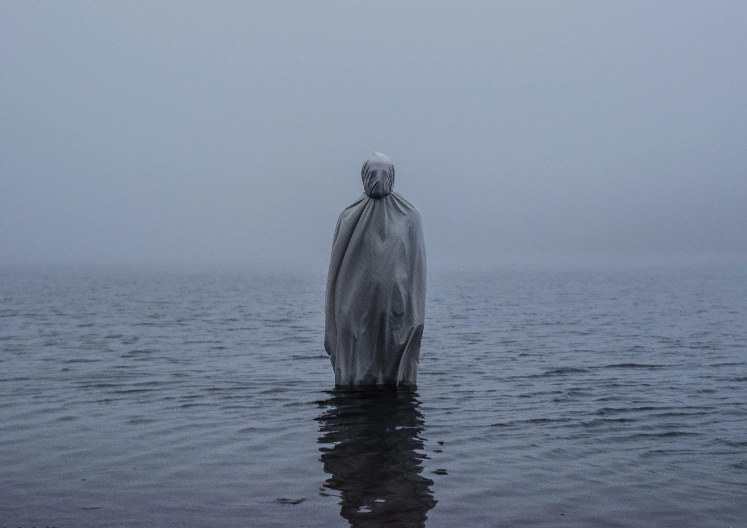 A person covered in a gray raincoat standing in calm water, facing away, with a foggy gray sky above.