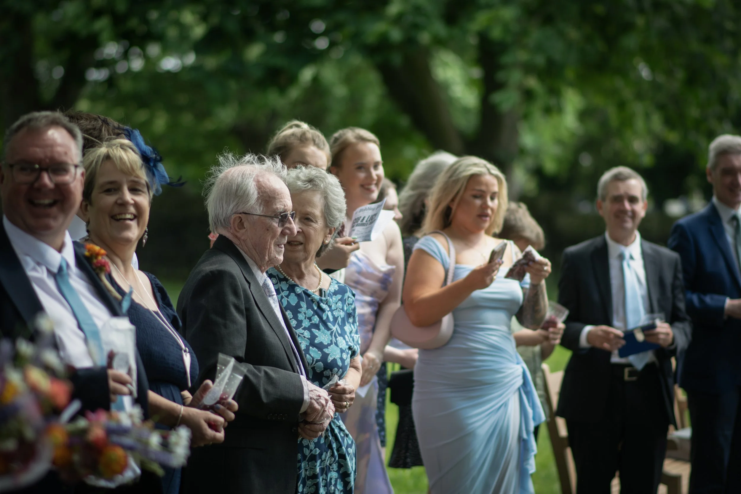 A group of people dressed in formal attire outdoors, likely celebrating a wedding, with some holding flowers and others reading programs or cards, and lush green trees in the background.